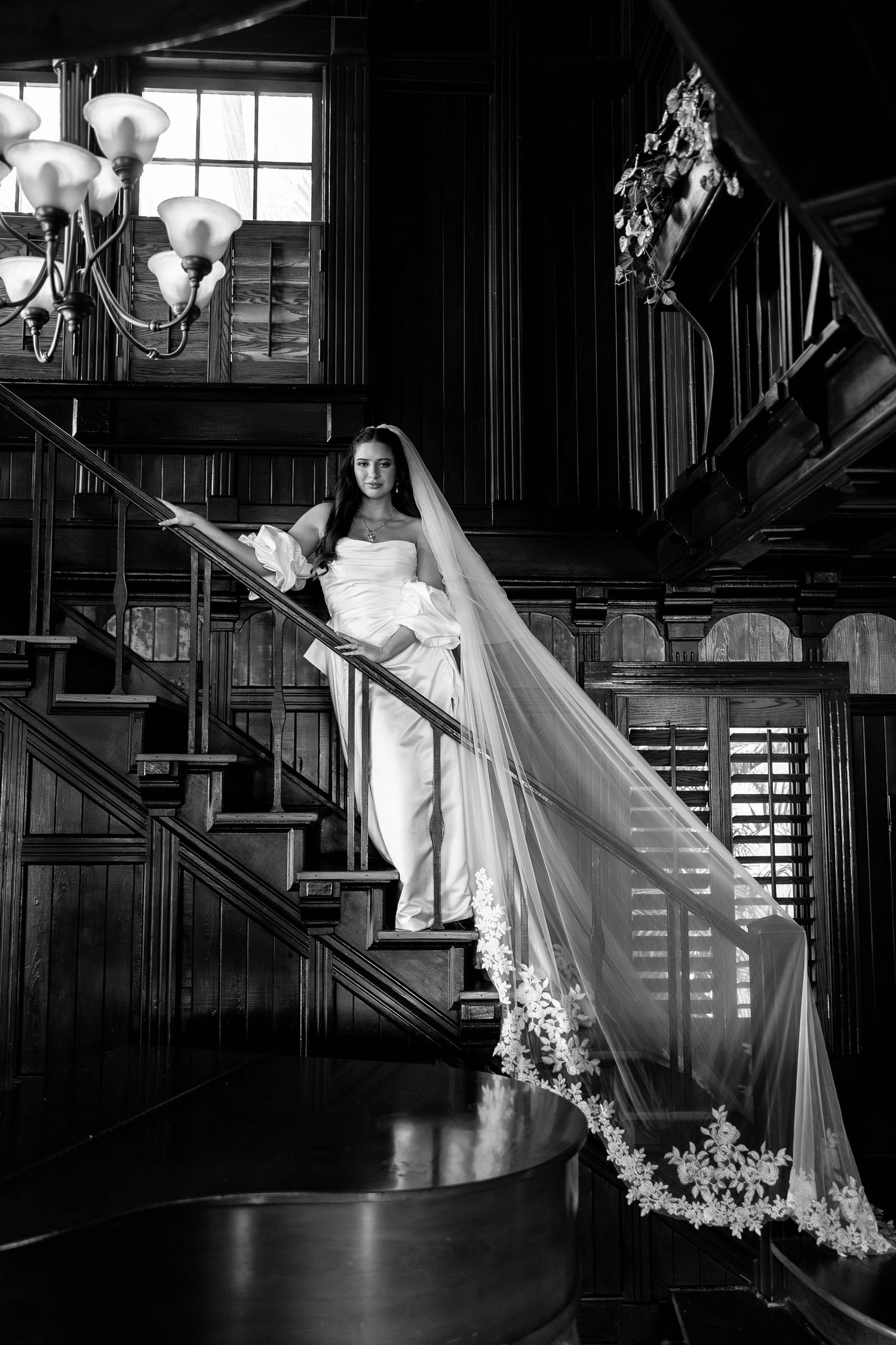 A bride in a flowing white gown and veil poses elegantly on a dark wooden staircase in dramatic black and white lighting.
