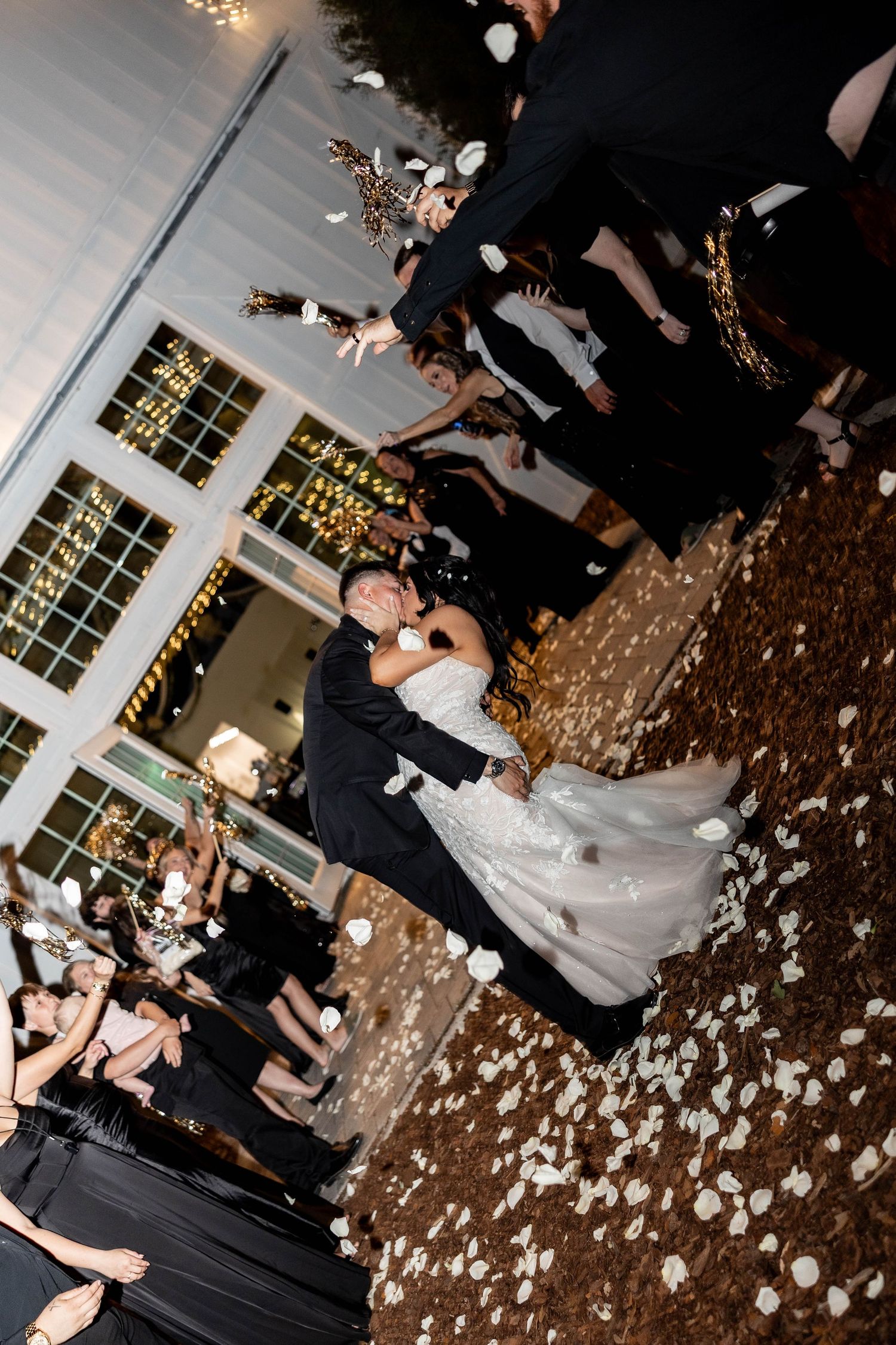 Bride and groom share a kiss as guests toss white rose petals during a romantic outdoor wedding send-off at night.