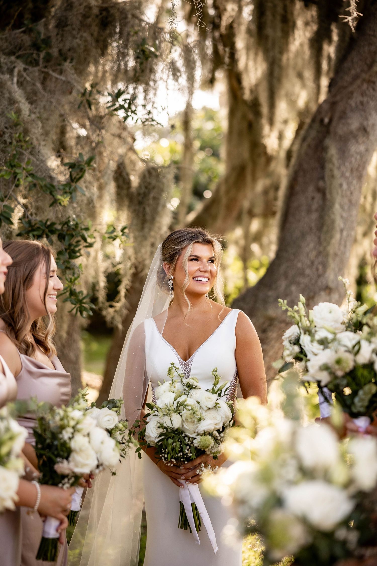 Wedding party members in white dresses gather under moss-draped oak trees with floral bouquets at outdoor ceremony.