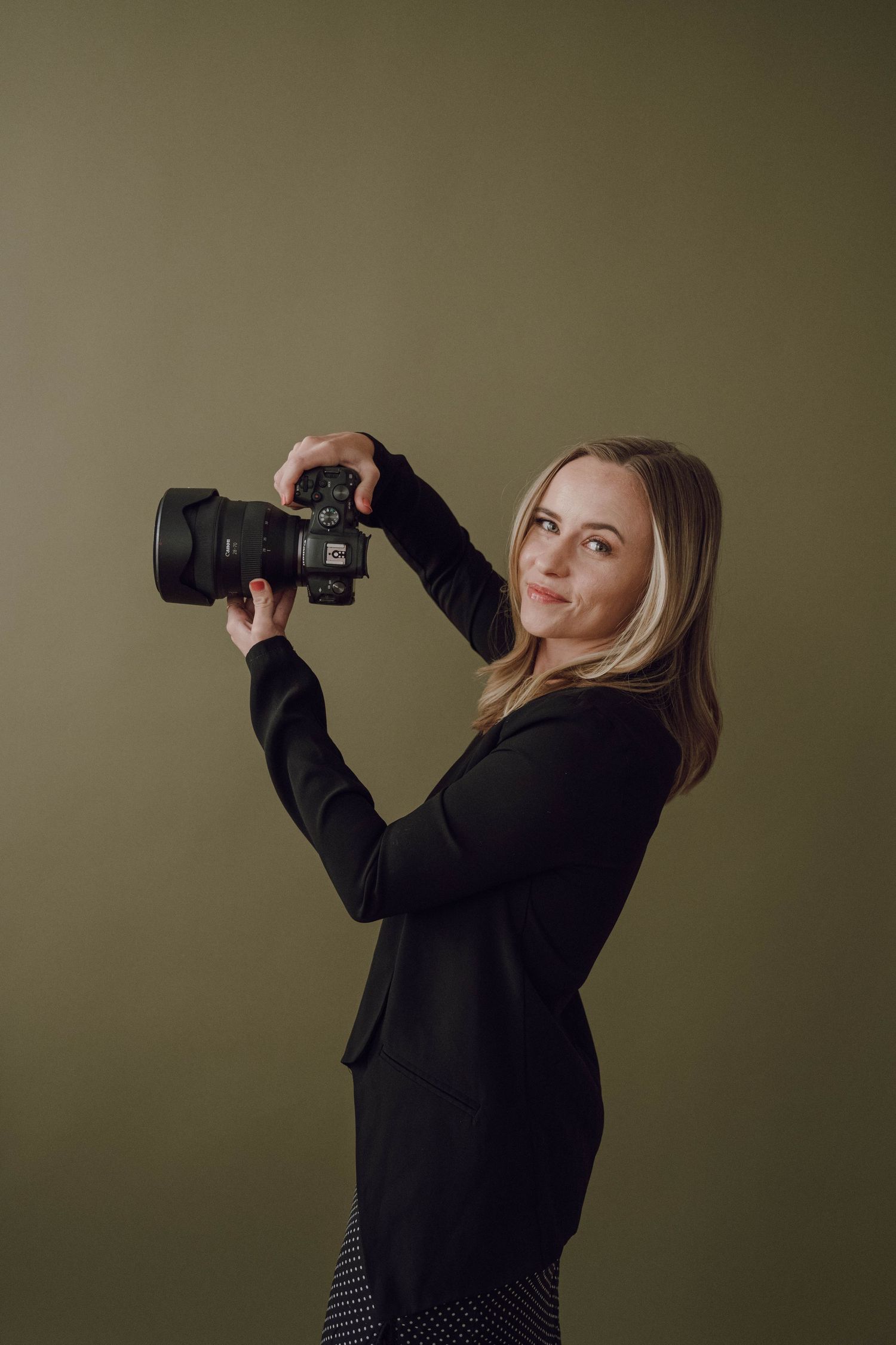 Professional photographer in black sweater poses with DSLR camera against olive green backdrop.
