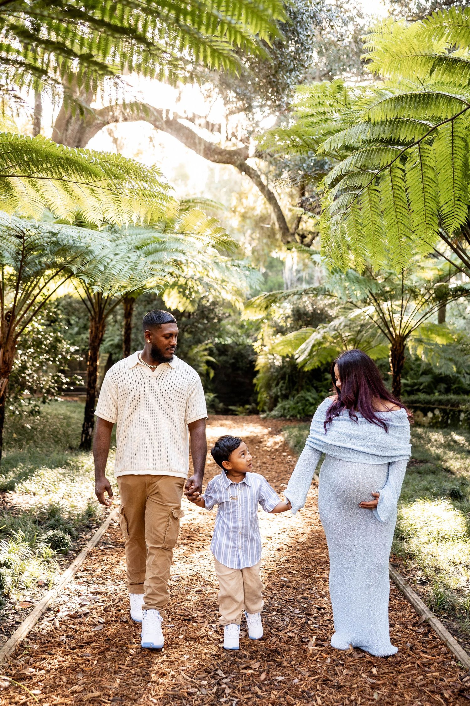 A family of three walks hand-in-hand through a lush garden, with a pregnant mother in a gray dress.