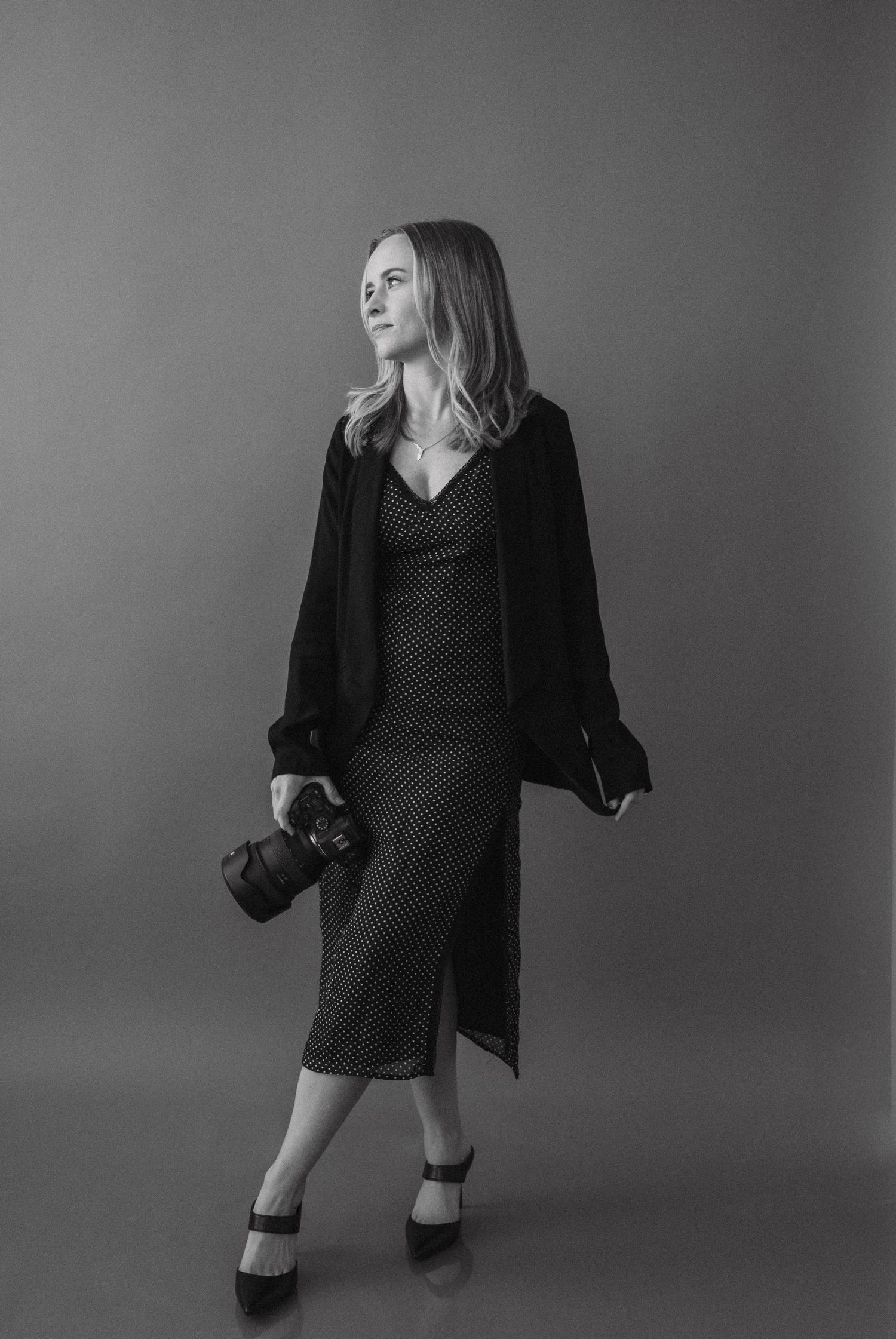 Black and white portrait photo of someone in a polka dot dress and cardigan holding a camera against gray backdrop.