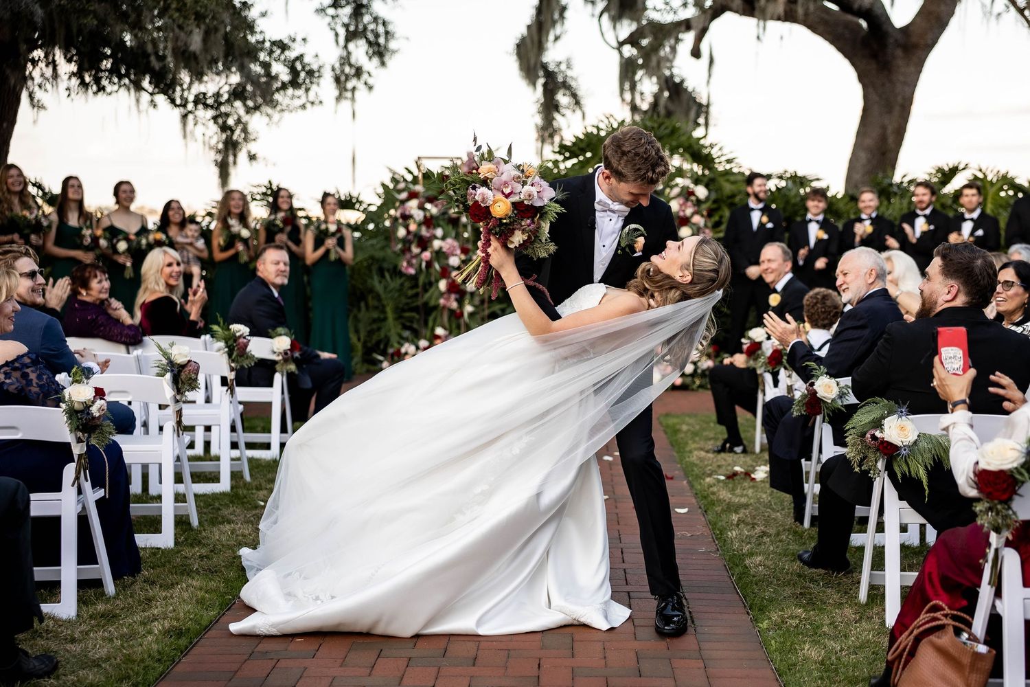 Wedding guests watch as a bride's dress flows in the wind beneath a large oak tree during an outdoor ceremony.