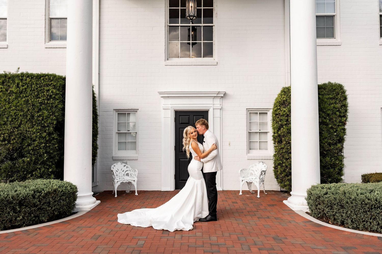 A series of white architectural columns and doorways with manicured hedges along a curved brick pathway.