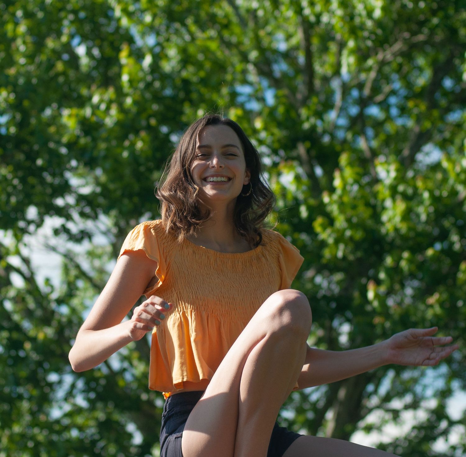Sarah, the photographer, sits and smiles on a sunny day, vivid green leaves of a tree all behind her