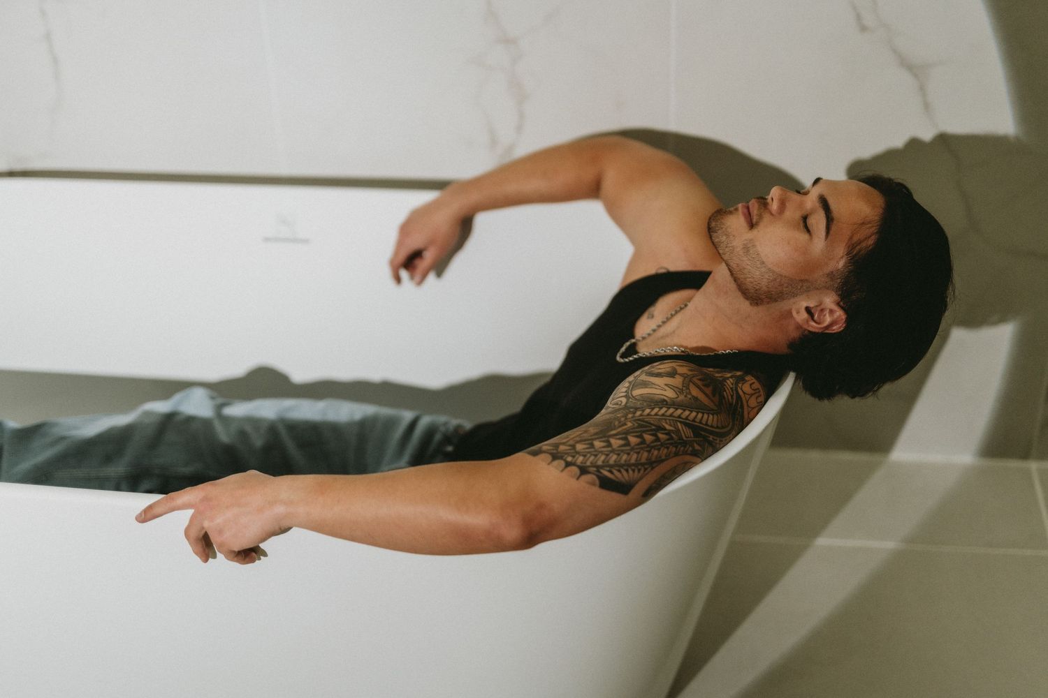 Man with tribal arm tattoo lying back in freestanding bathtub wearing black tank top and jeans in marble bathroom.