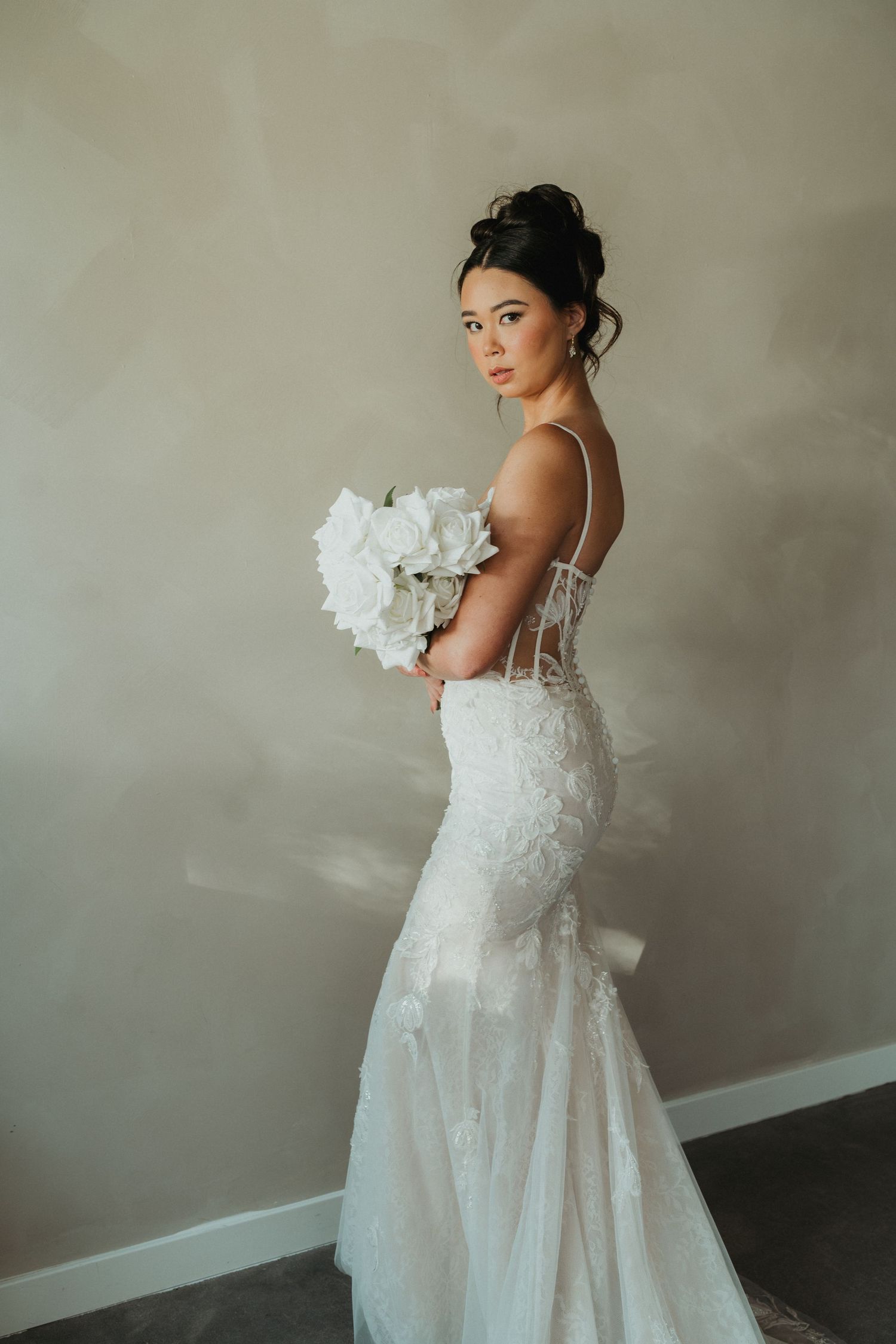 Beautiful bride in backless lace mermaid wedding dress holding white bouquet against beige wall.