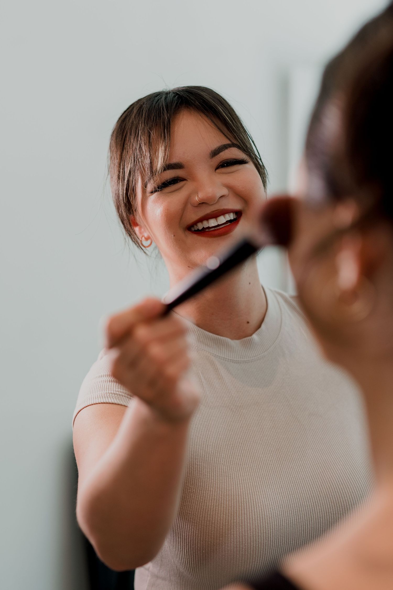 A smiling young woman in a white top laughs while a makeup artist applies blush with a brush.