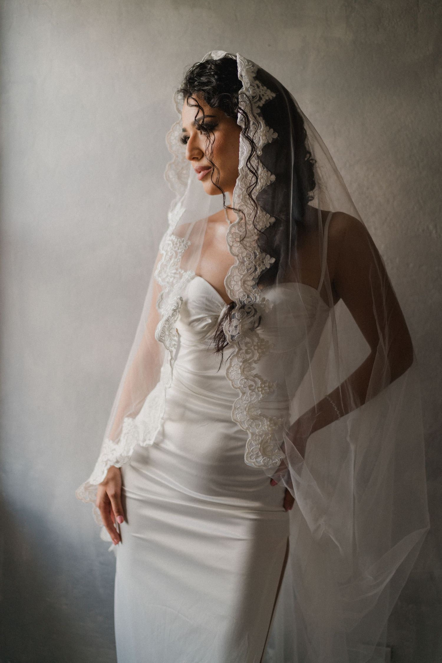 A bride in a fitted white strapless gown with a lace-trimmed veil poses gracefully against a gray wall.