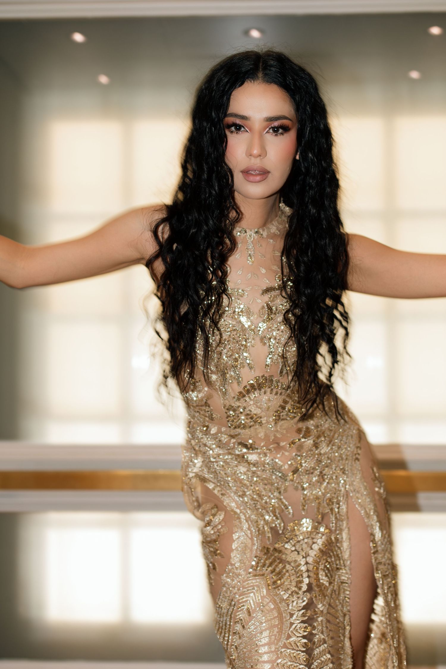 Woman in gold sequin gown with long curly black hair poses elegantly indoors.