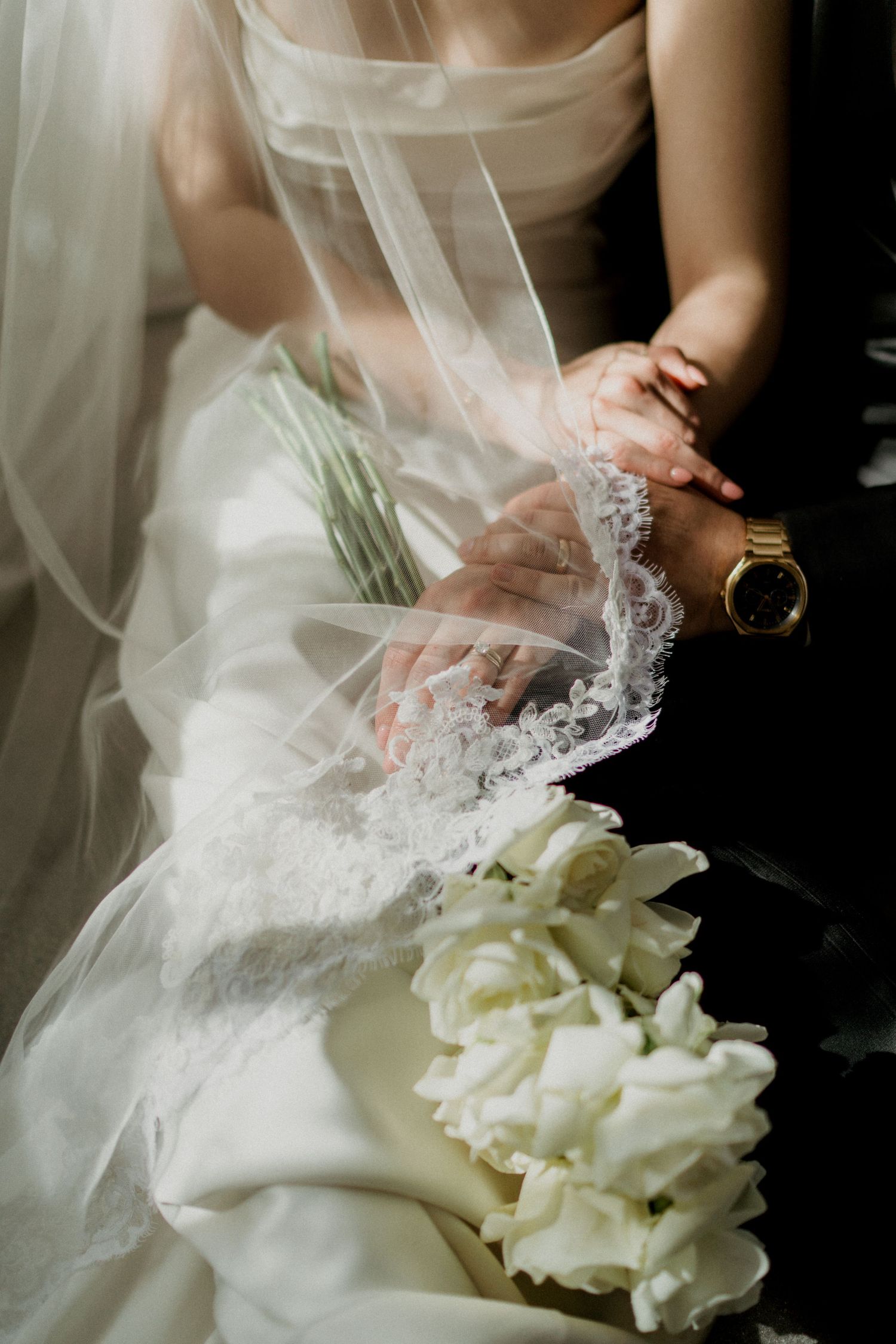 Close-up of bride holding white rose bouquet with lace veil draped over hands and gold watch.