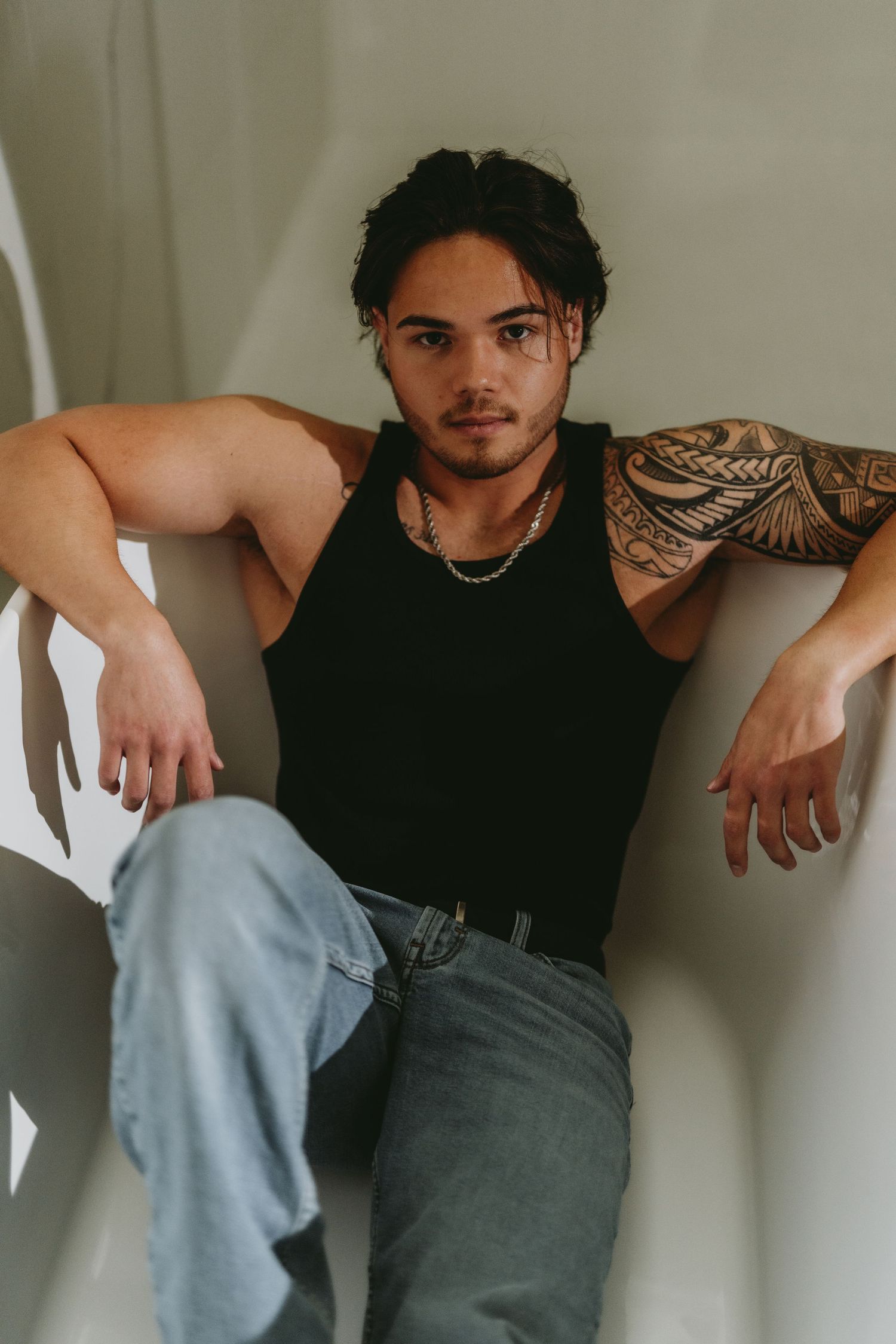 Young man with Polynesian sleeve tattoo in black tank top and jeans sitting against white freestanding bathtub.