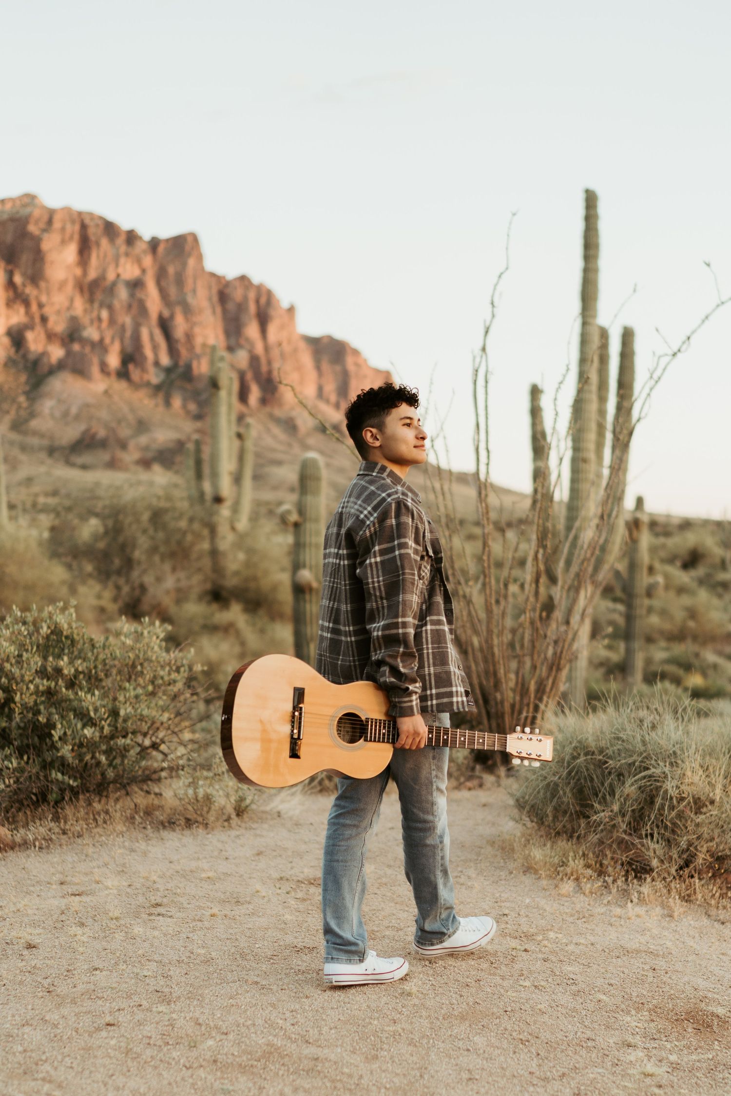 Young musician holding acoustic guitar looking up in scenic desert with tall saguaro cacti at dusk.