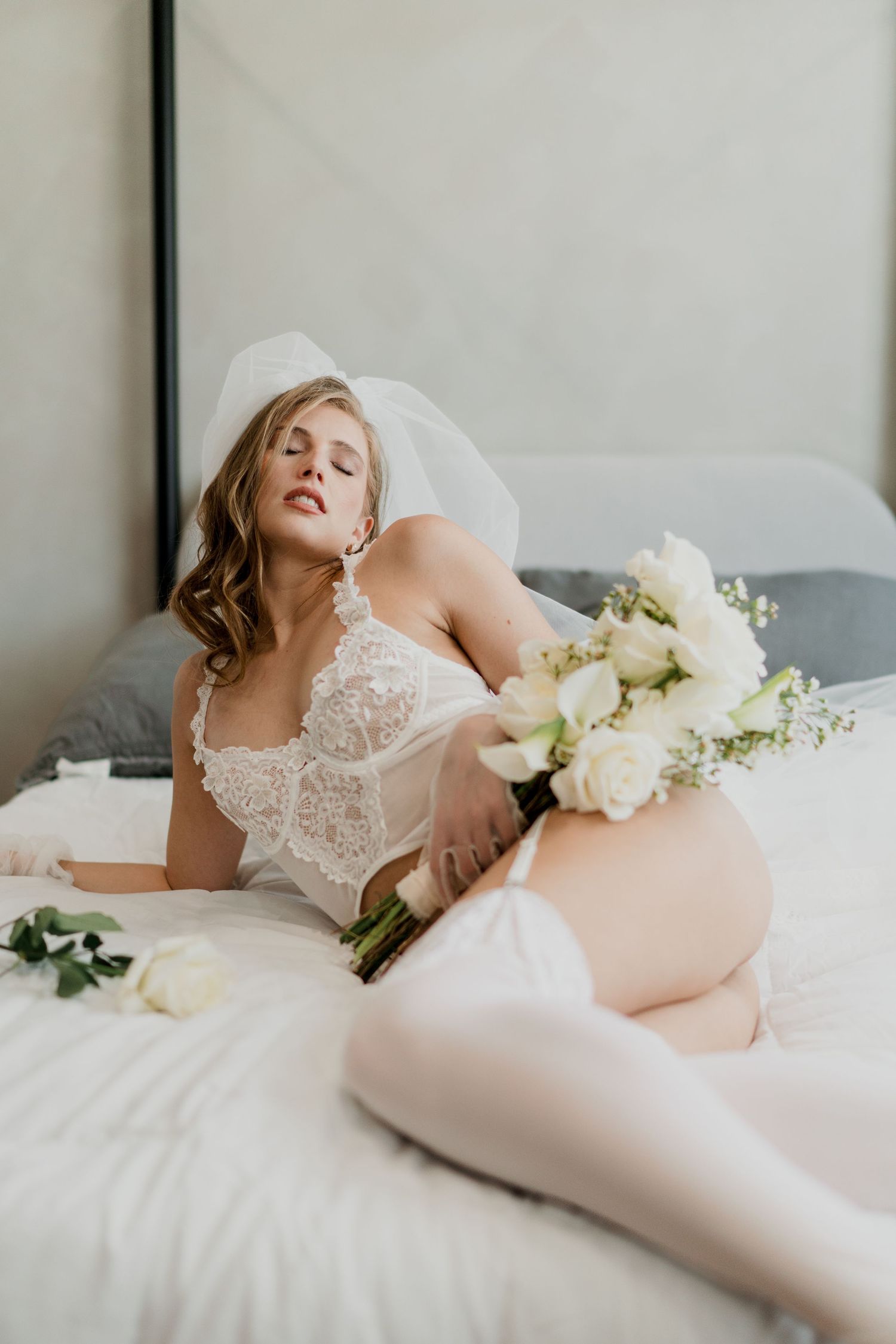 A bride in white lace lingerie and veil sits on a bed holding a bouquet of white roses.