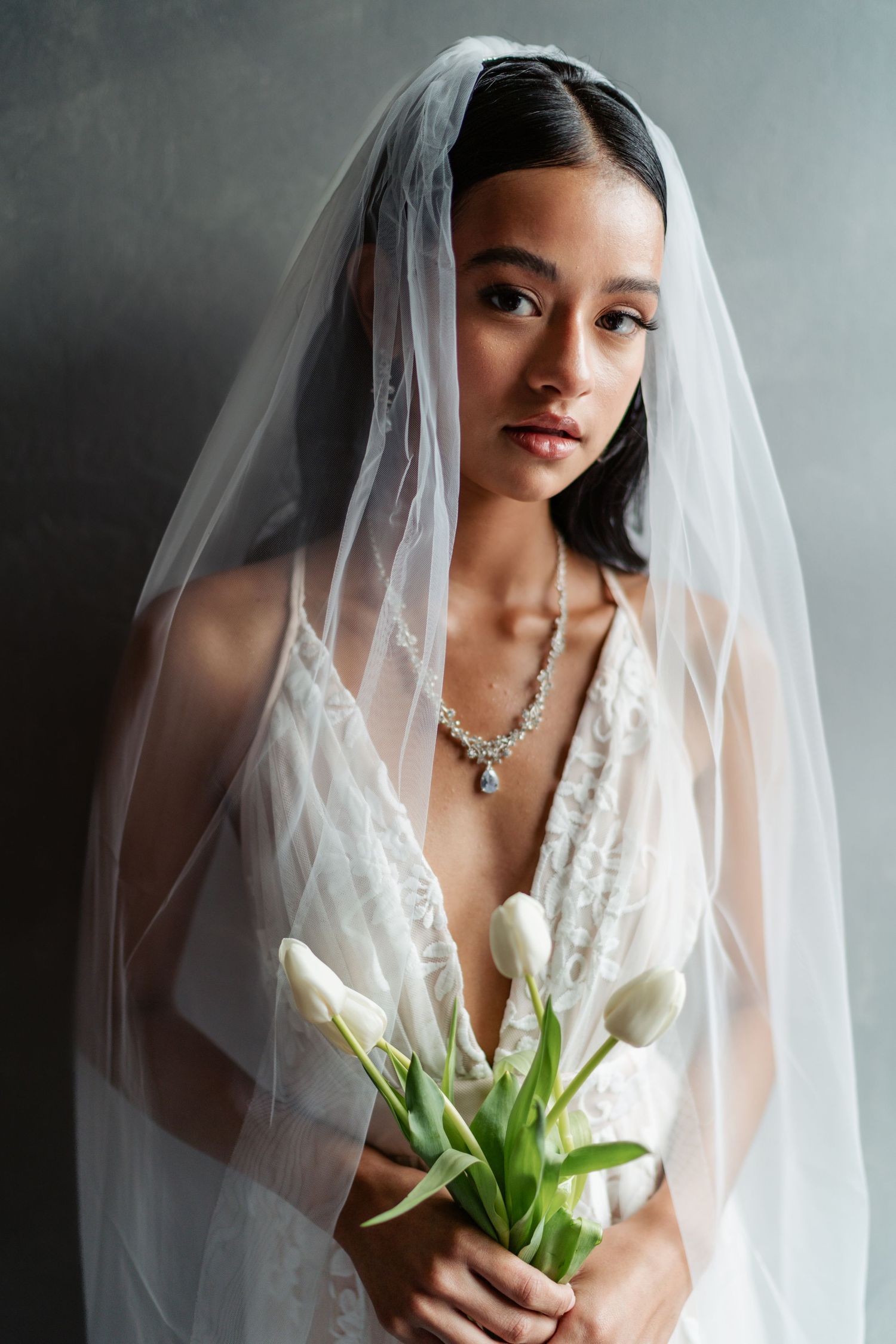 A bride in a white lace dress and veil holds white tulips while looking directly at the camera.