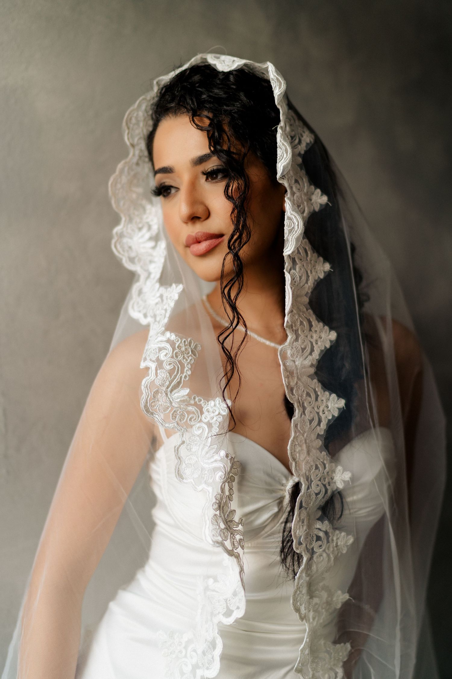 A bride in a white strapless dress with an ornate lace veil gazes softly at the camera.