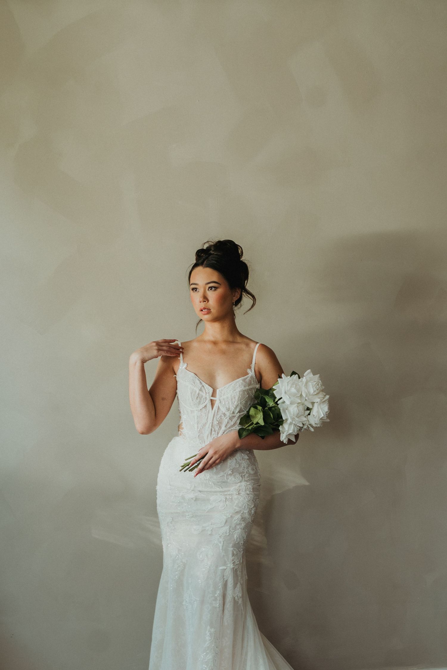 Elegant bride in lace mermaid wedding gown holding white peony bouquet against neutral wall.