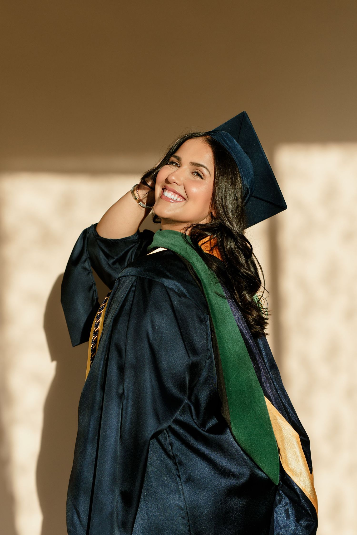 Smiling young woman in navy graduation cap and gown with green and gold sash.