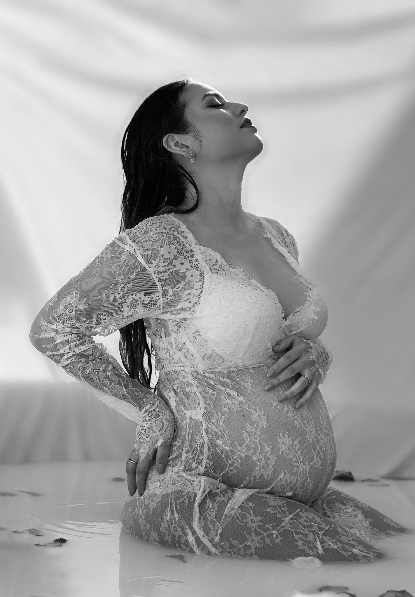 Black and white artistic maternity photo of pregnant woman in lace dress posing gracefully on floor.