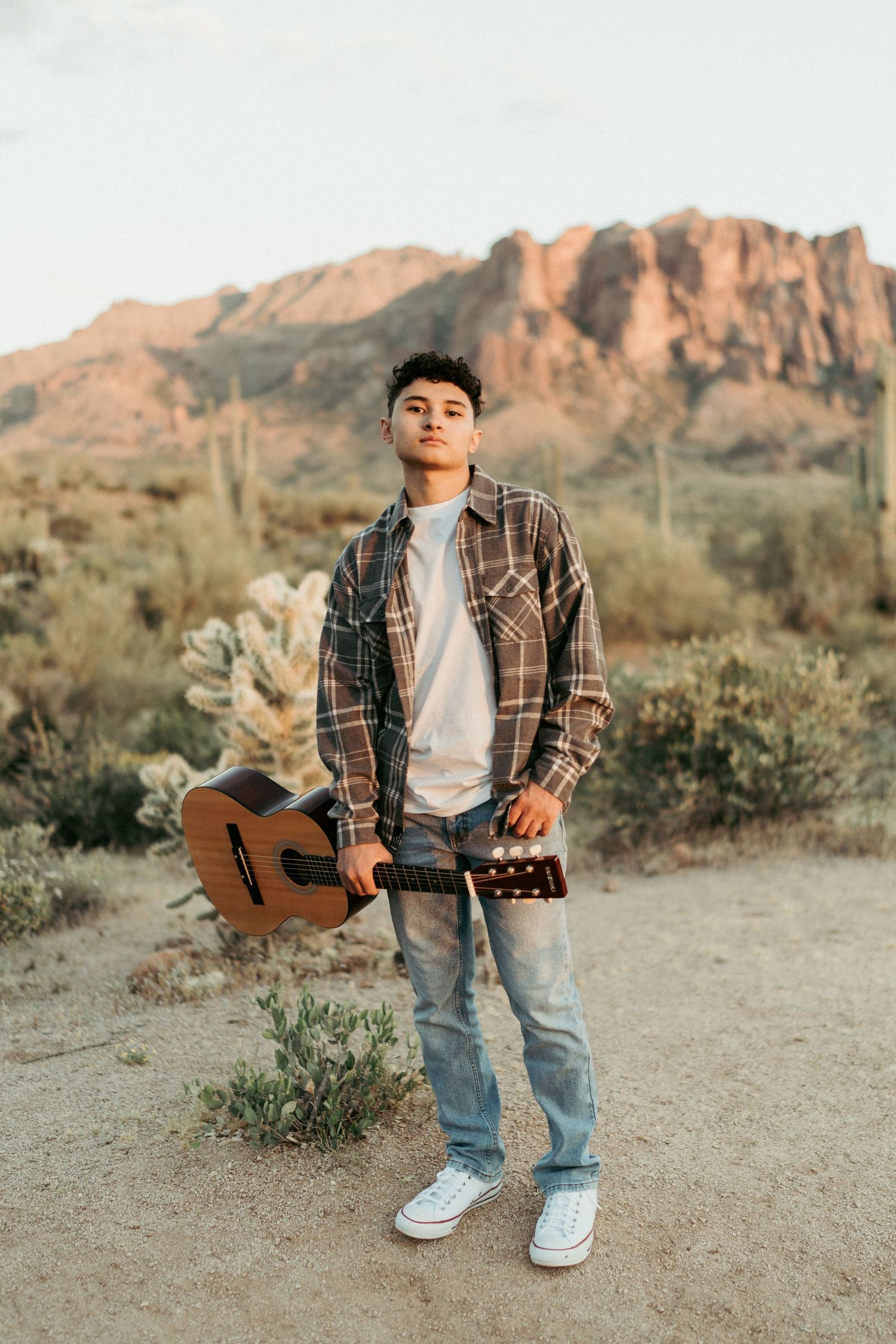 Young man holding acoustic guitar standing in Arizona desert landscape with saguaro cacti at sunset.