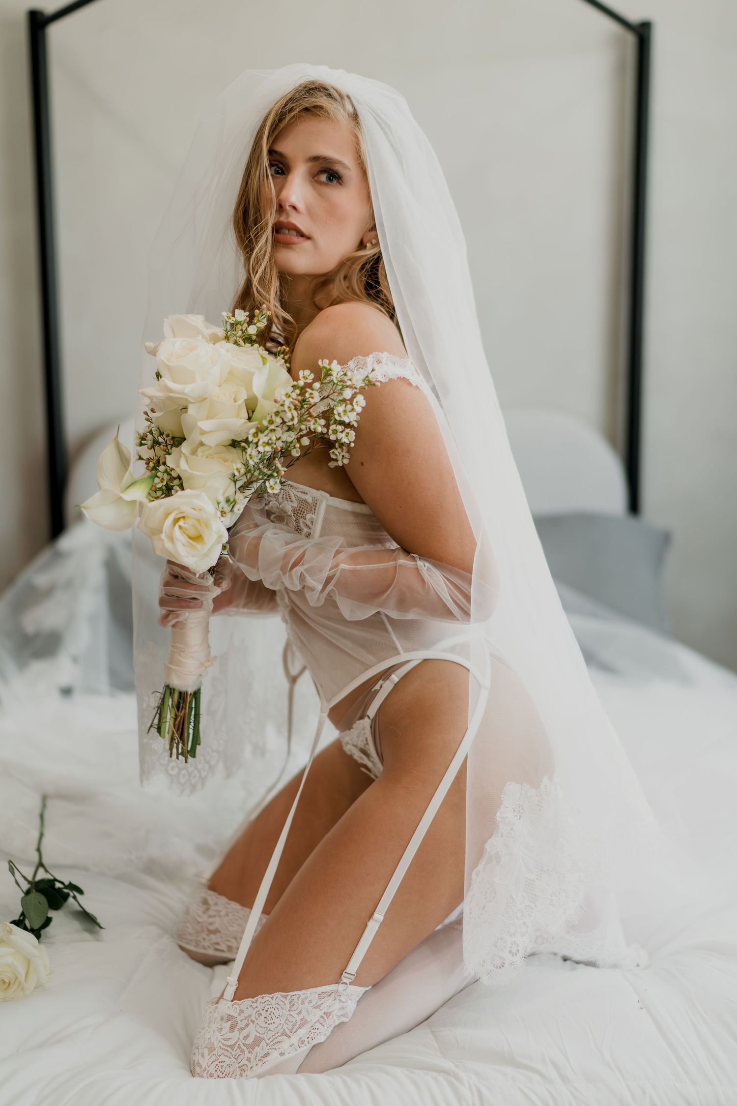 A bride kneeling on a bed in sheer white lingerie and veil holds a white floral bouquet.