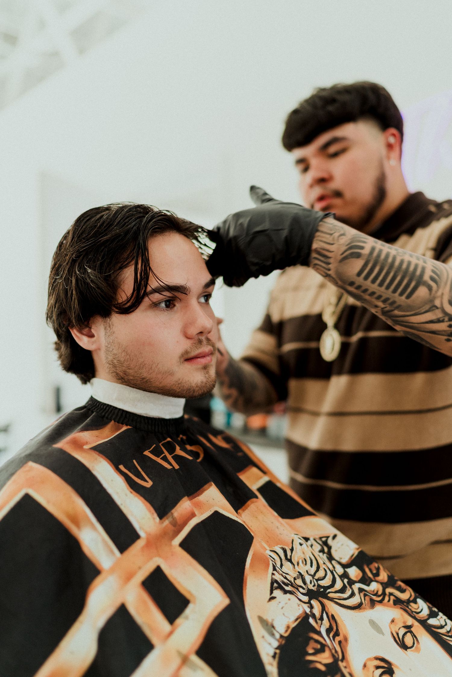 Young man getting a haircut at a barbershop by a tattooed barber wearing black gloves.