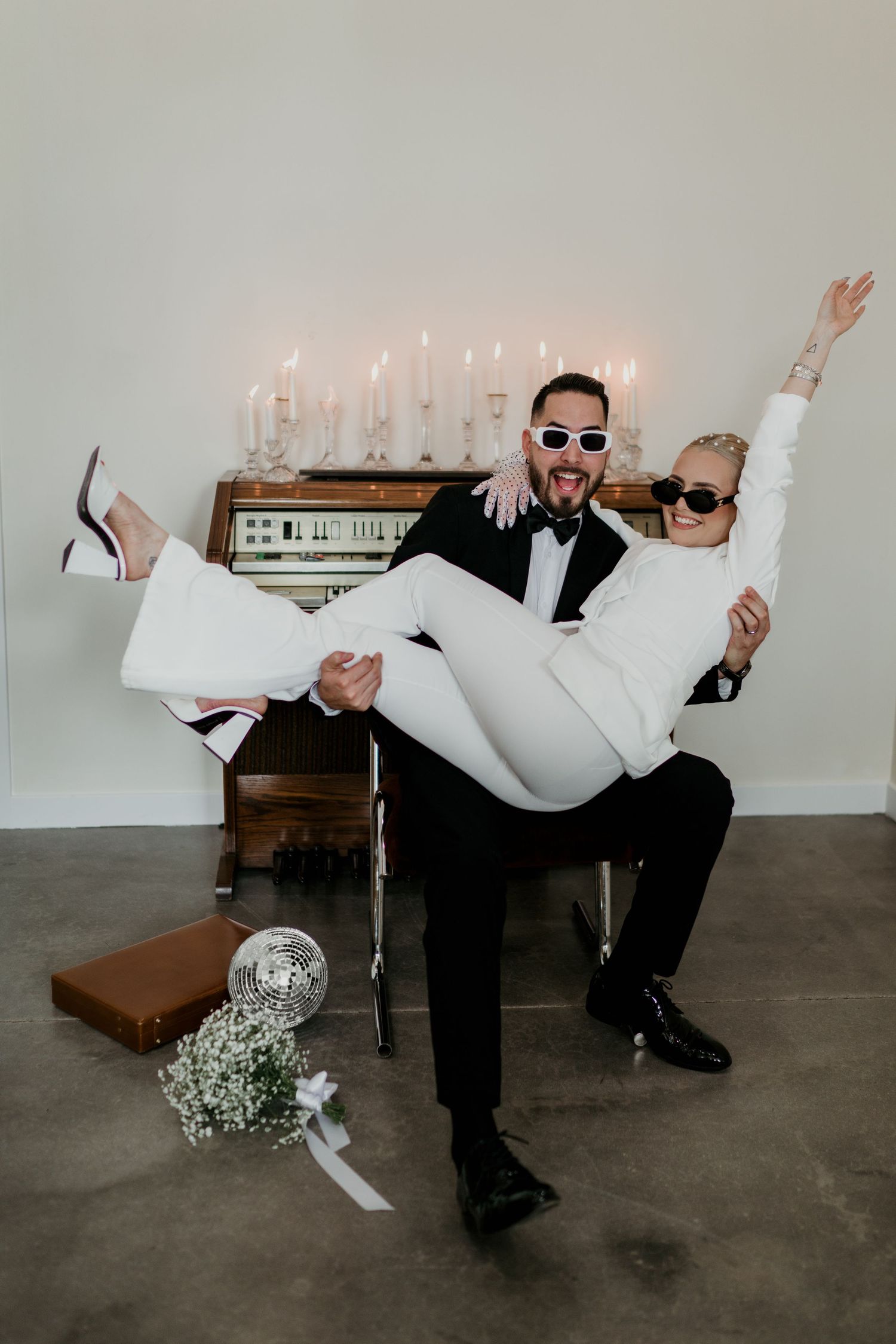 Couple in wedding attire posing playfully with bride sitting on groom's lap in front of candelabra and vintage piano.