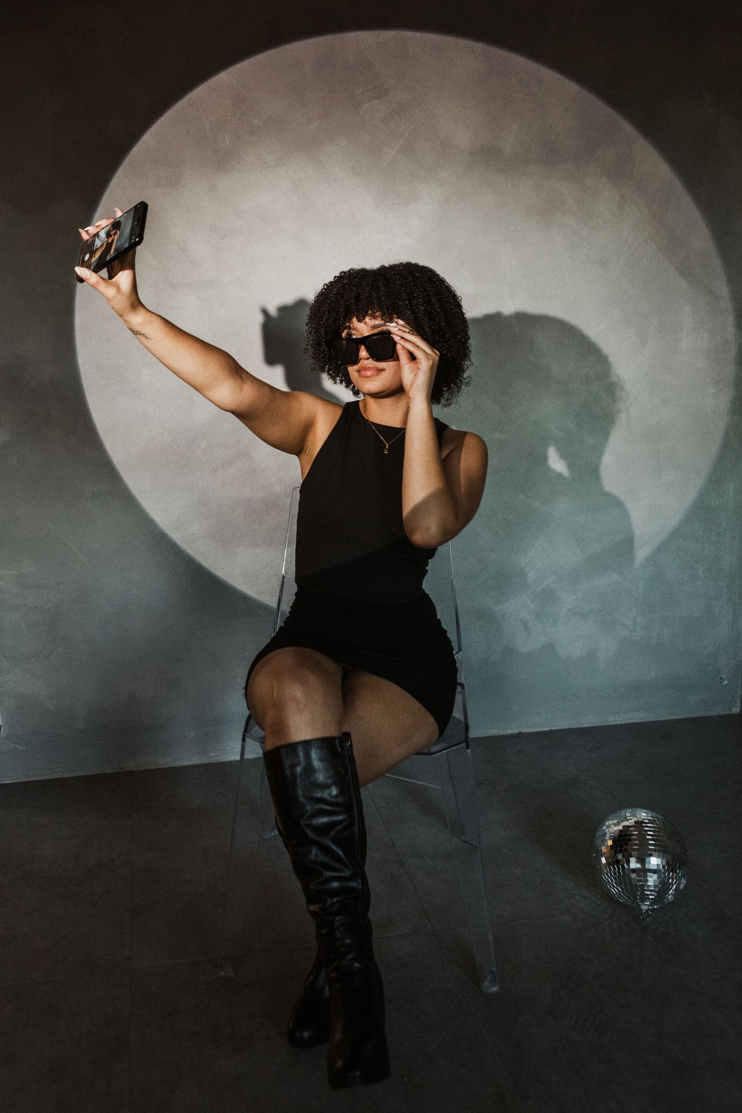 A woman in a black dress and sunglasses takes a selfie while sitting on a chair with a disco ball nearby.