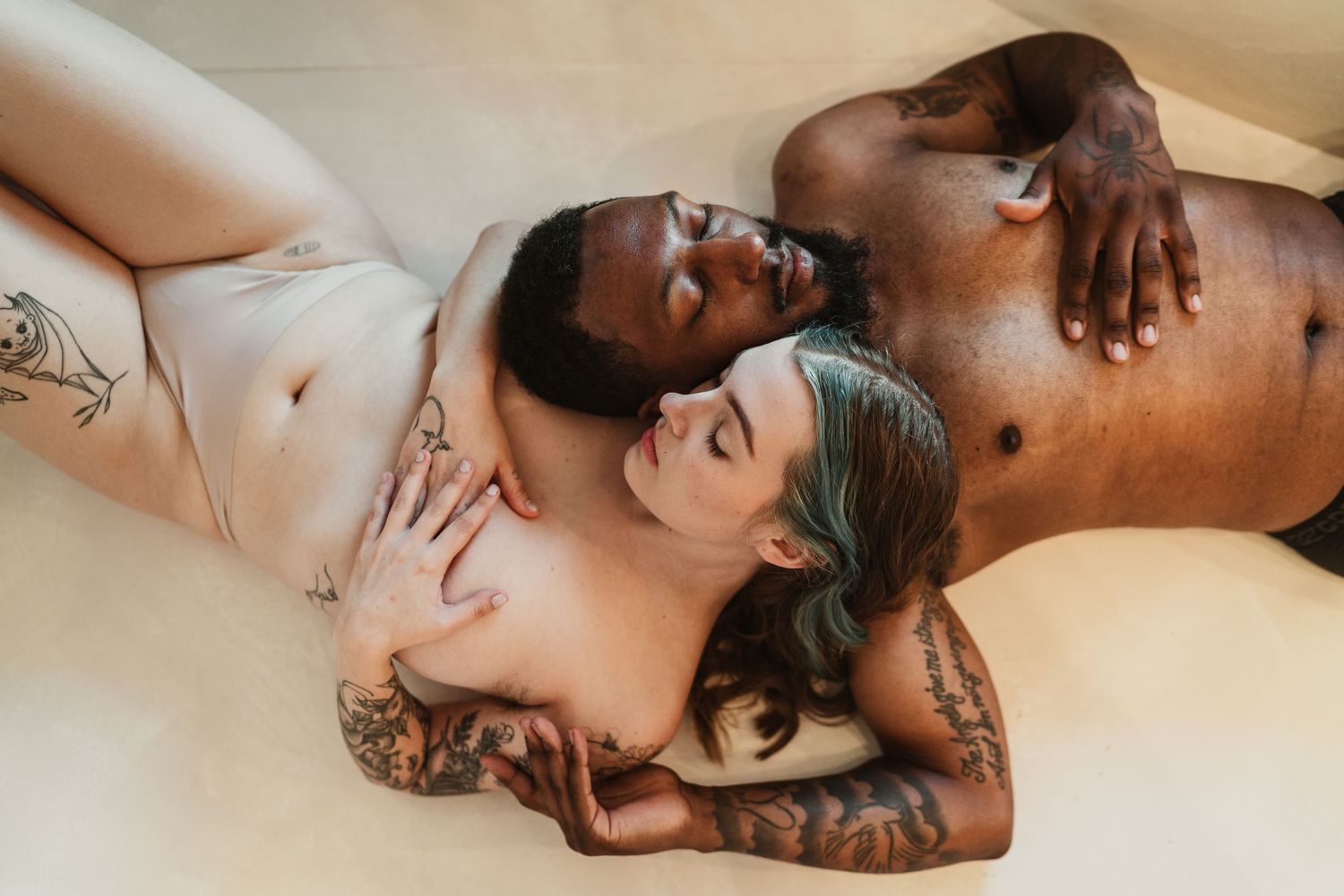 Tattooed interracial couple lying together intimately on a light-colored surface in soft lighting.