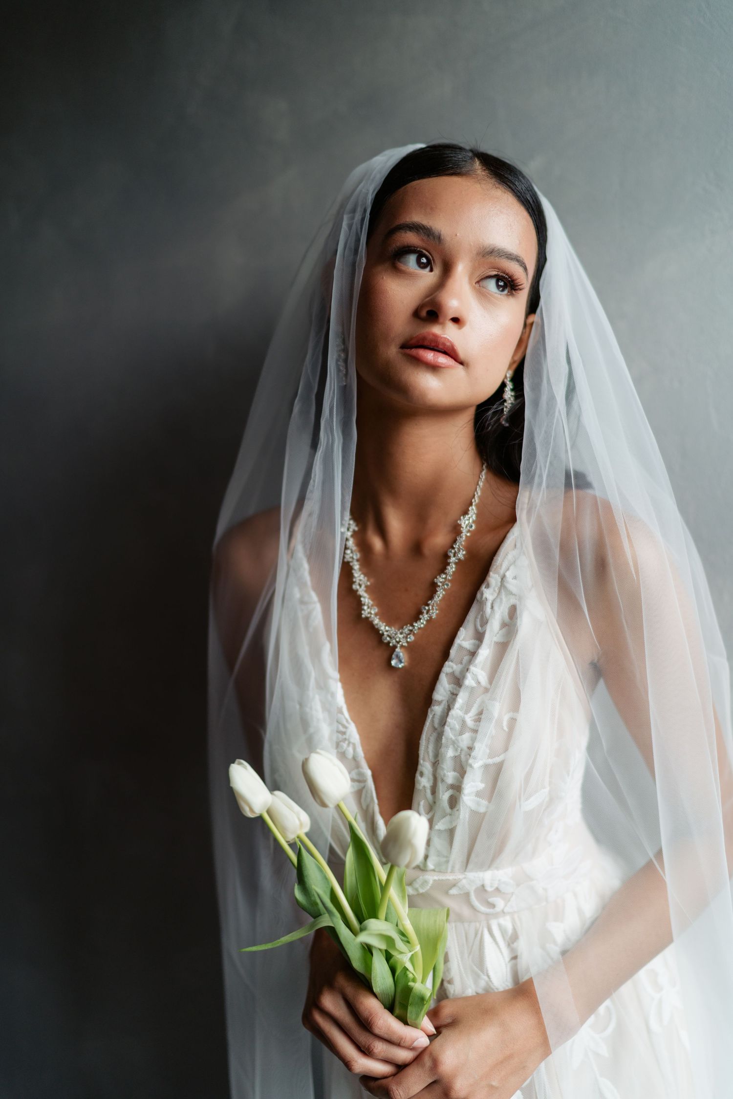 A bride in a white lace gown and veil holds white tulips while gazing upward against a gray background.