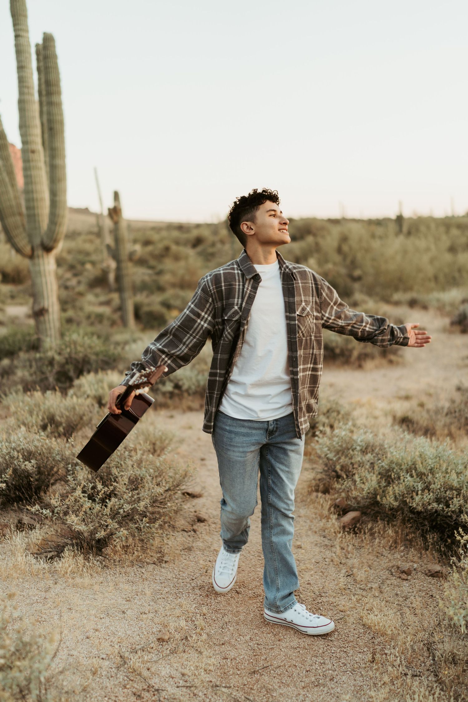 Joyful young man with outstretched arms holding guitar in desert landscape with saguaro cacti.