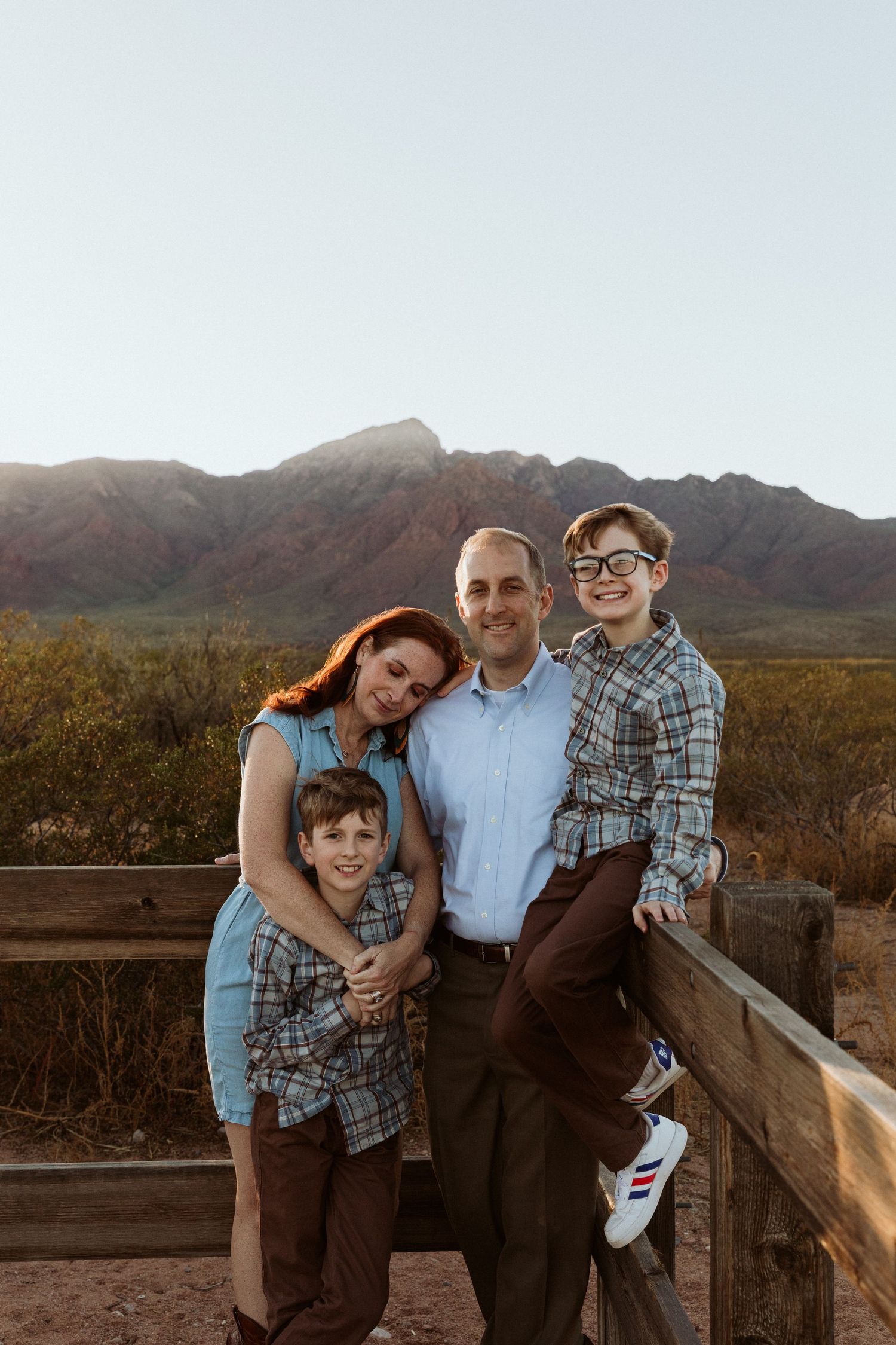 Family of four smiles together at wooden fence with mountain sunset backdrop.