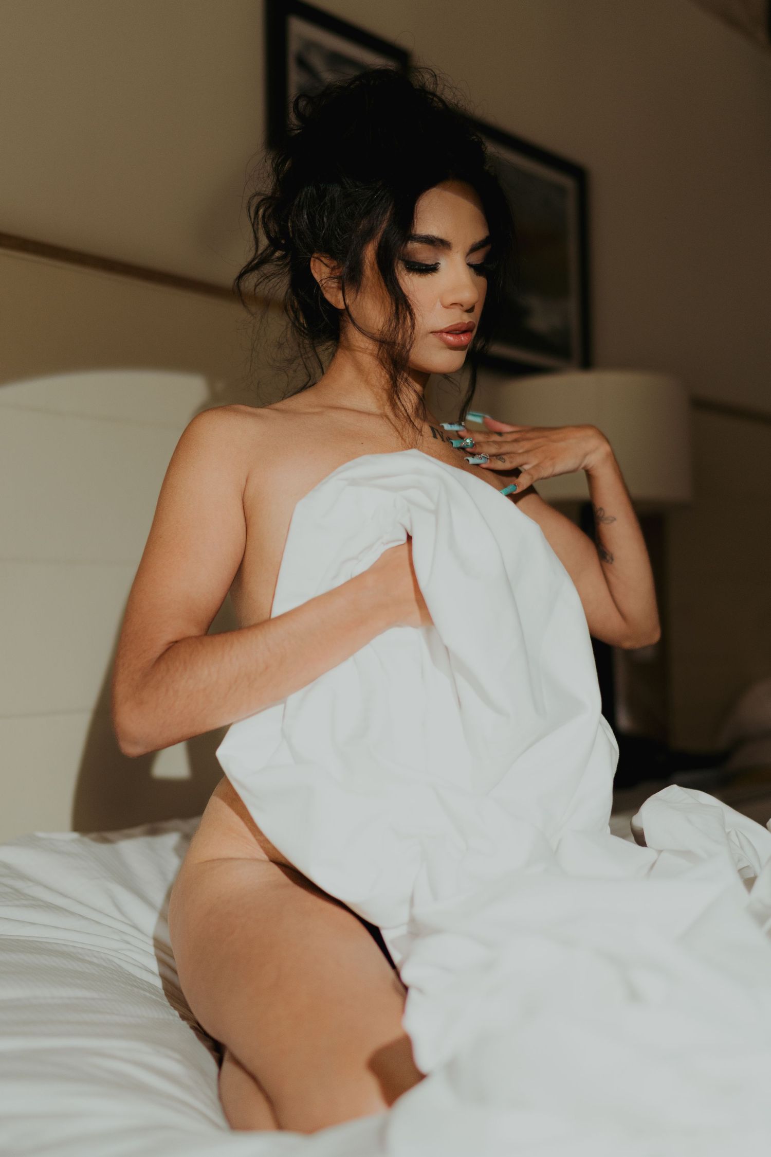 A woman wrapped in a white sheet sits on a bed wearing nude underwear with dark upswept hair.