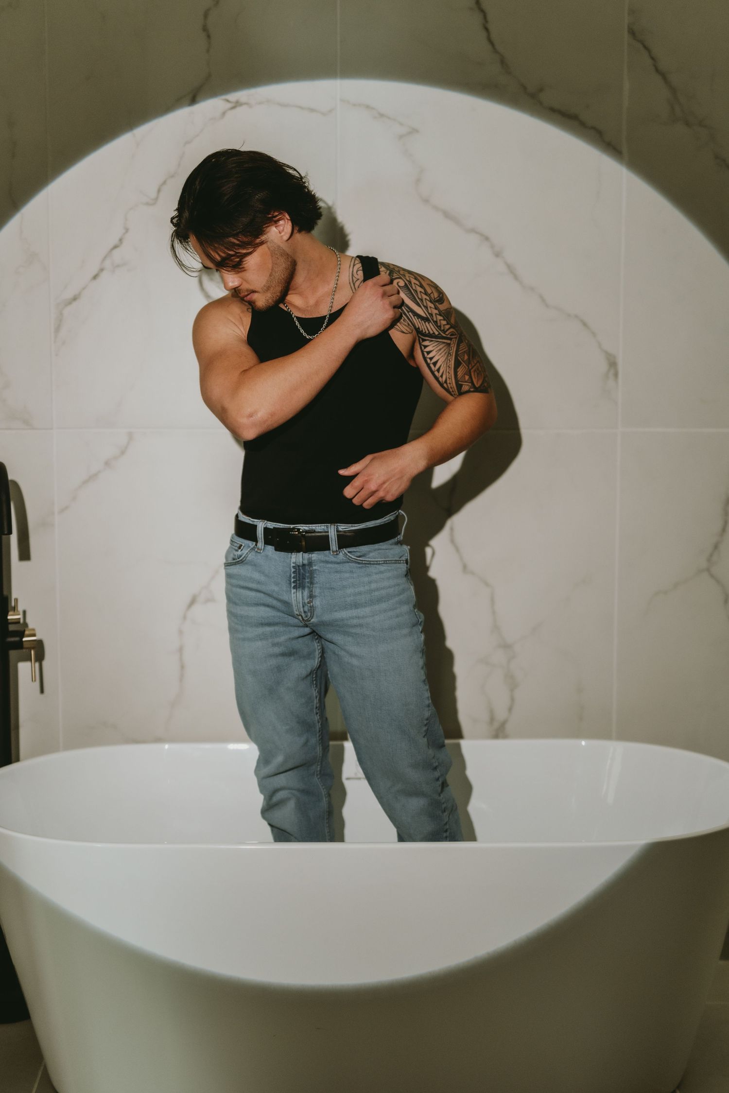 Muscular man with tribal arm tattoo in black tank top and jeans posing by freestanding bathtub in marble bathroom.