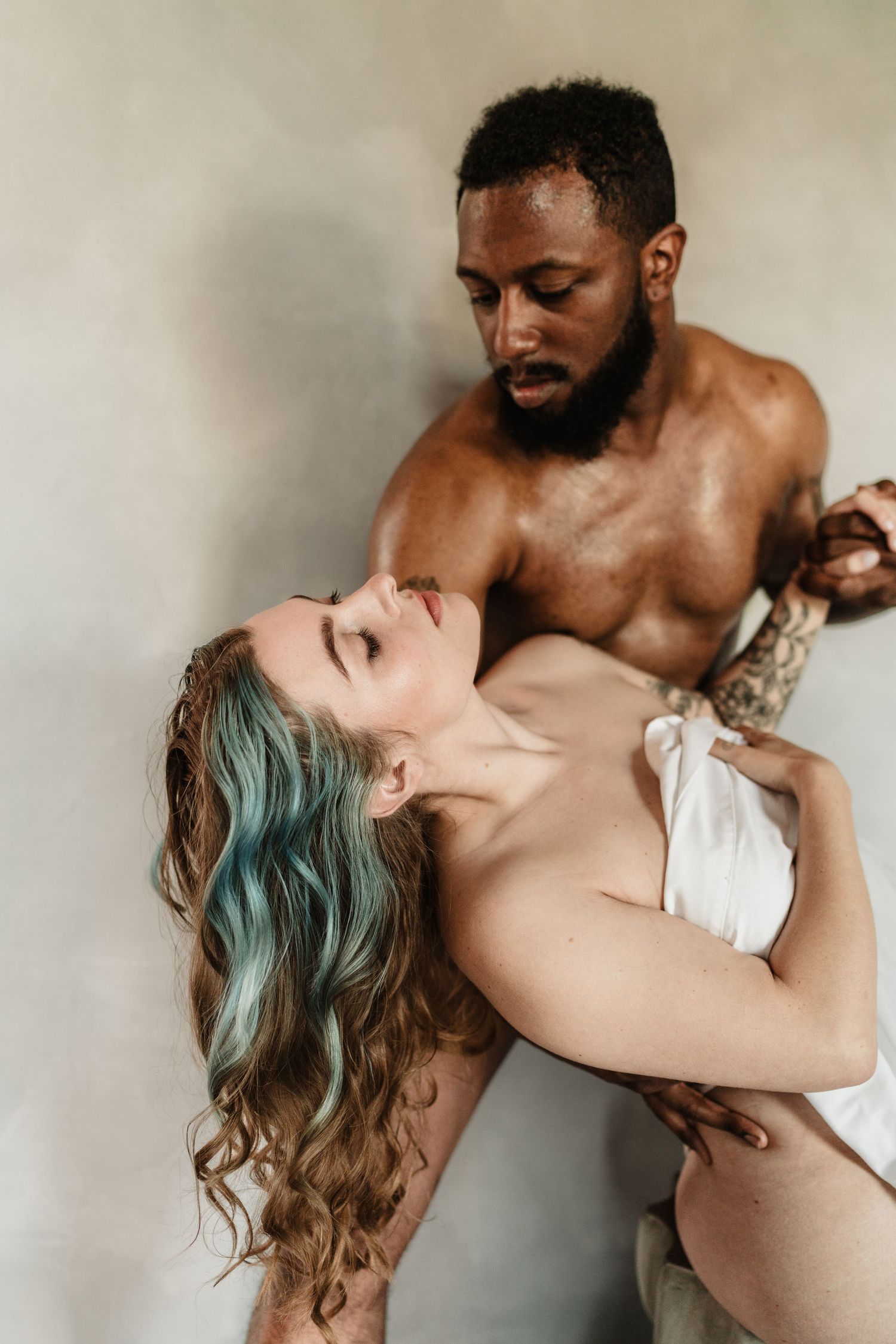 Shirtless tattooed Black man holds woman with blue-highlighted hair in an intimate studio portrait.