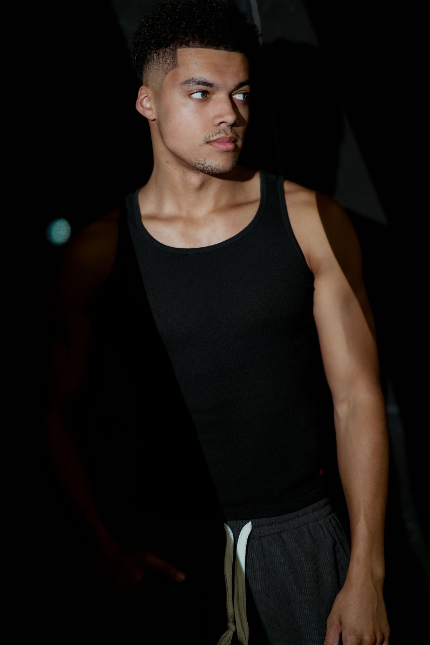 Athletic young man in black tank top and gray shorts posing in dramatic dark lighting.
