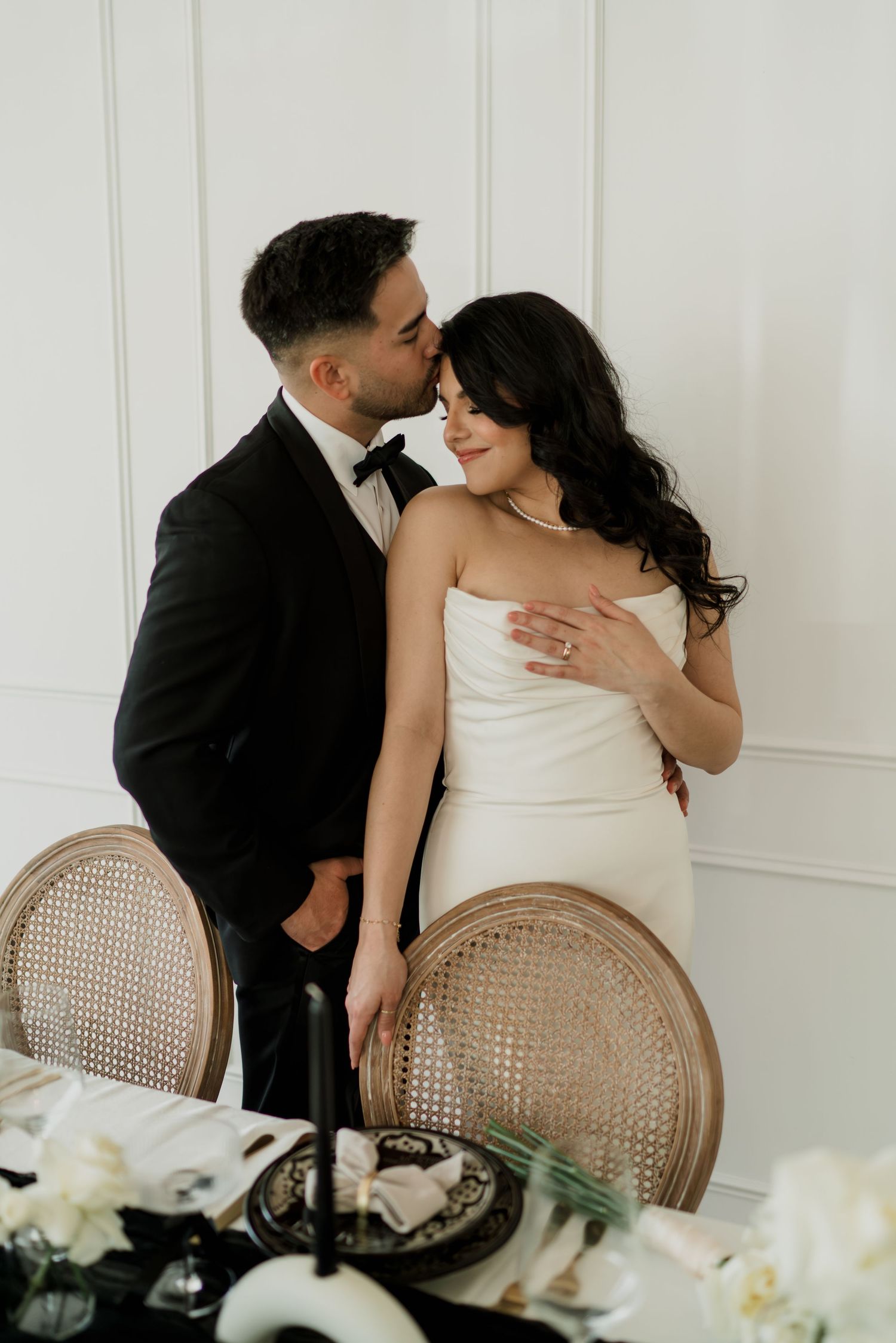 Groom in black tuxedo kisses bride in white strapless dress beside elegant wedding reception table.