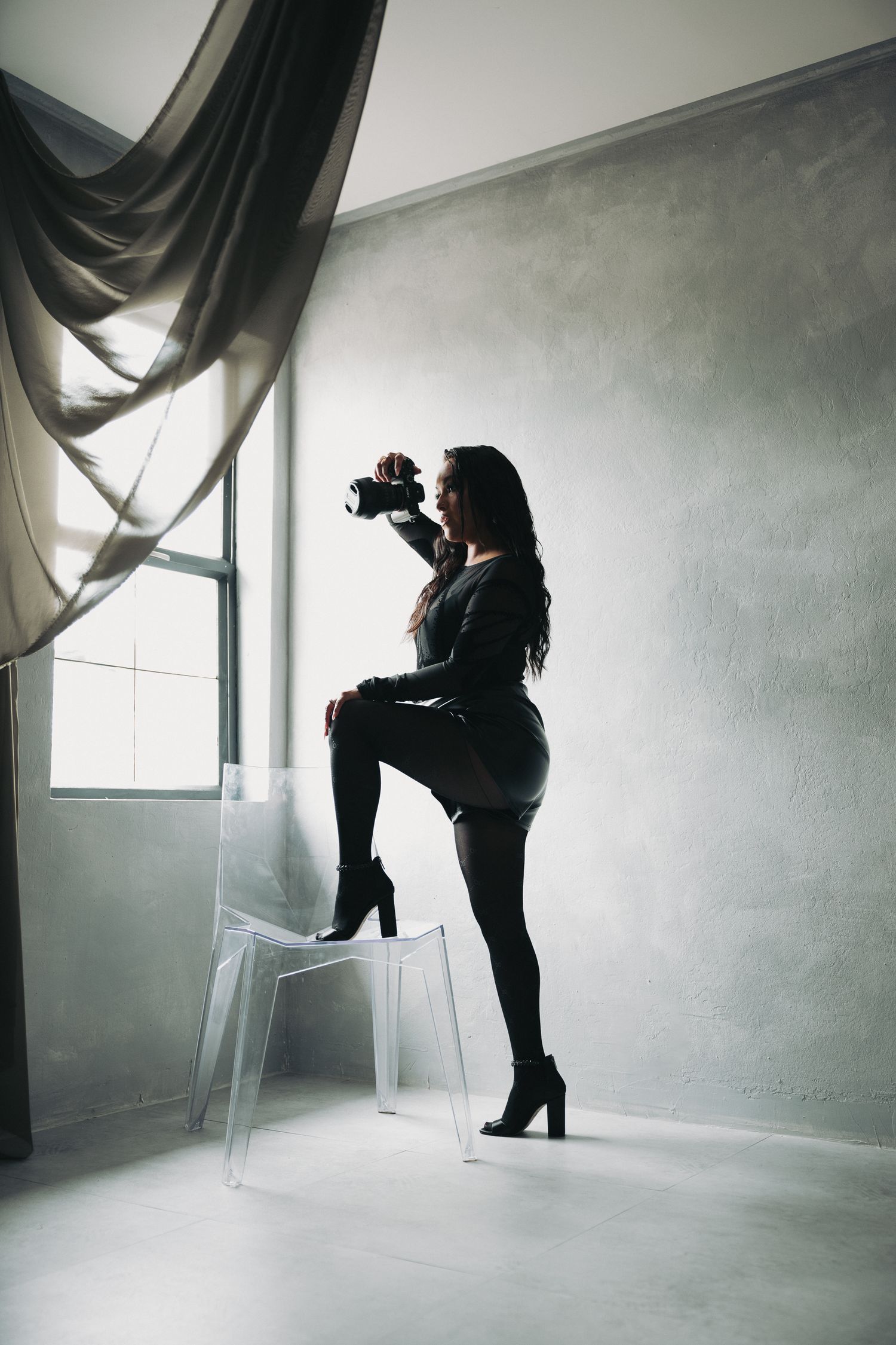 Silhouette of woman in black outfit photographing with DSLR camera while posing on chair near studio window.