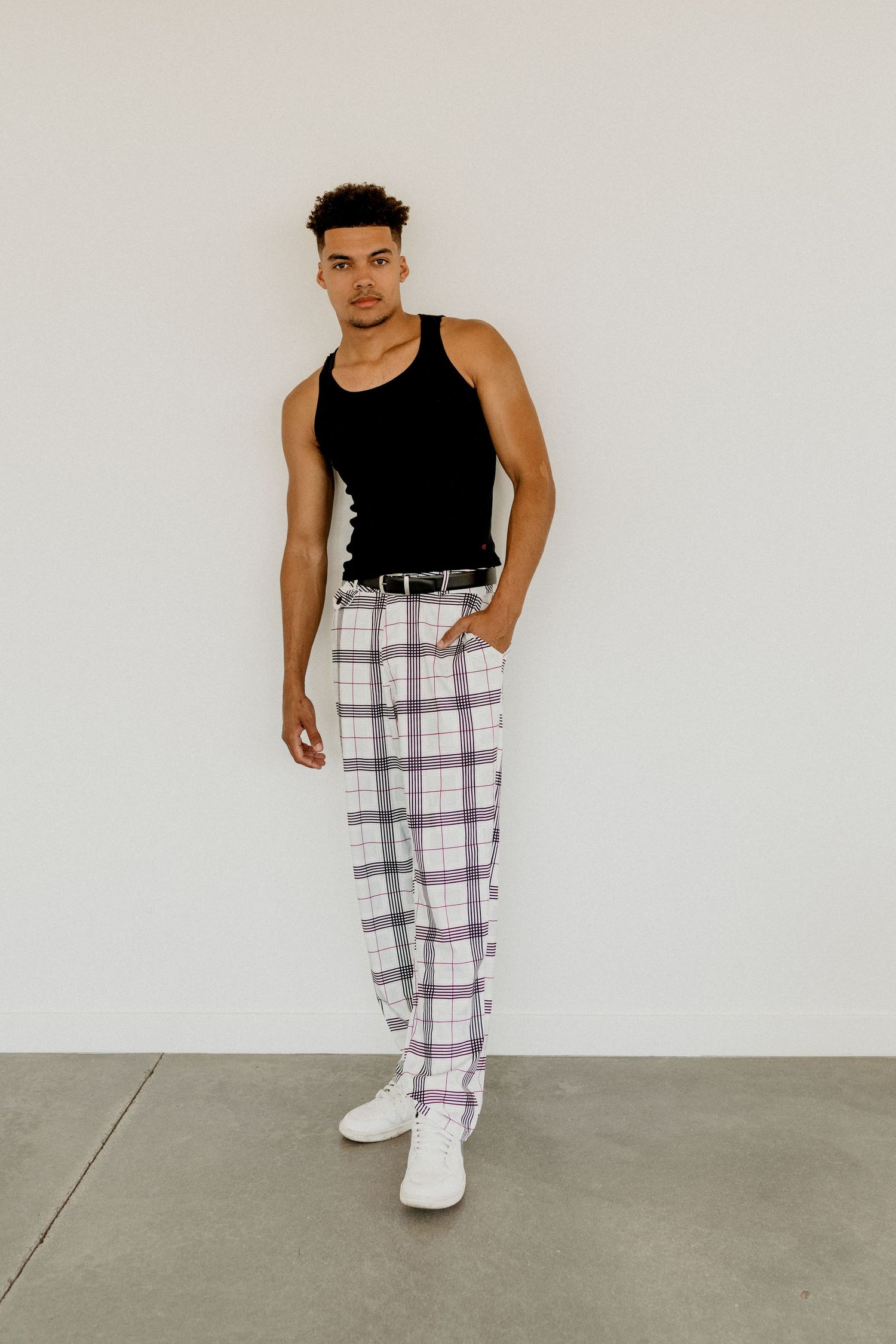 Stylish man in black tank top and plaid trousers posing against white wall.