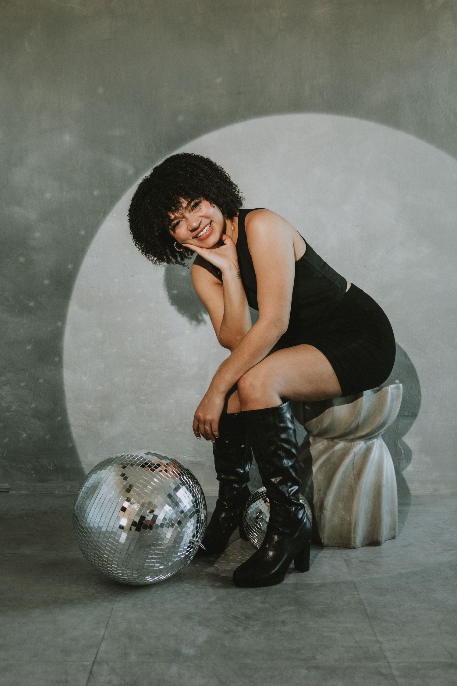 Smiling woman with curly hair in black dress poses on pedestal beside disco ball in studio.