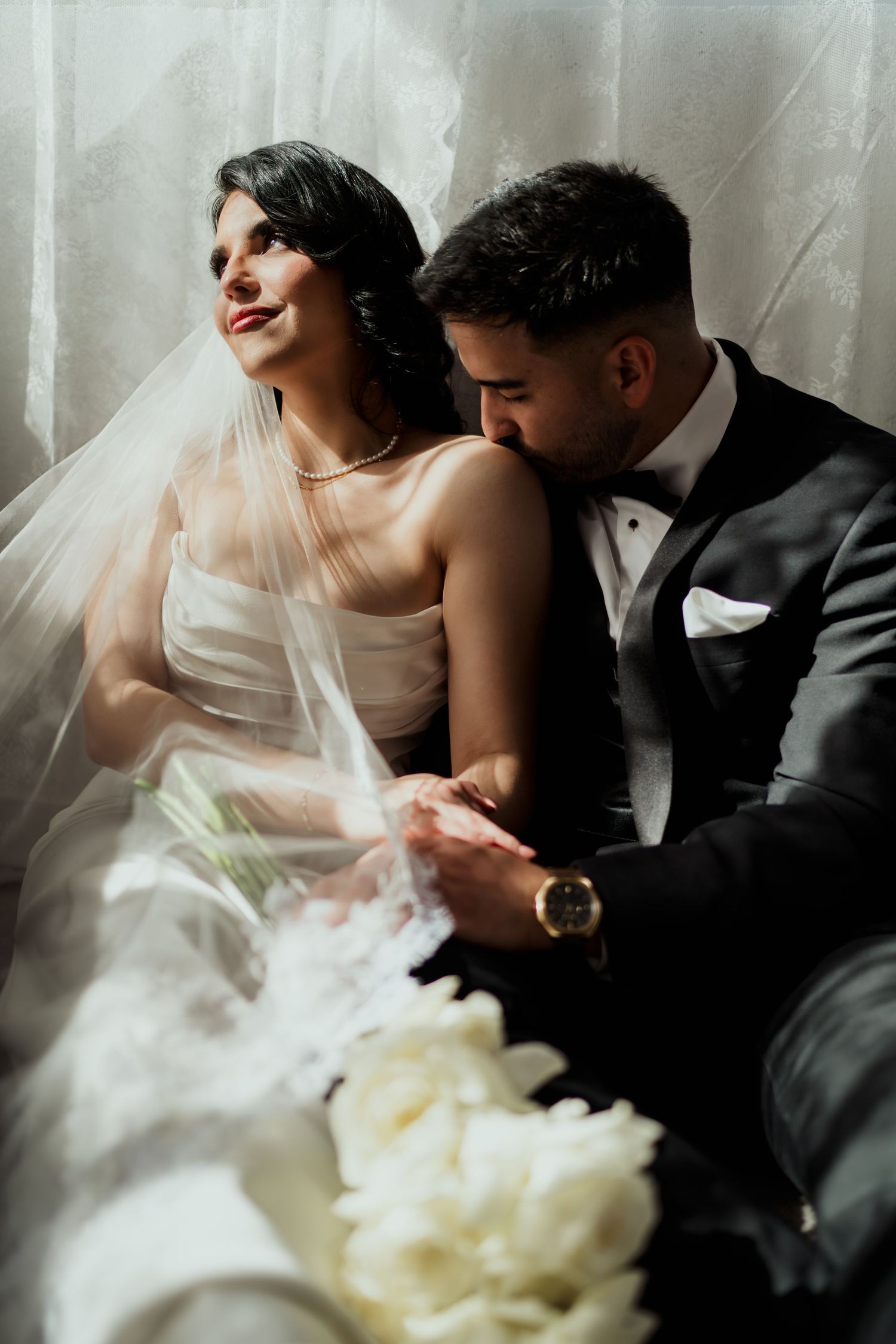 Bride in white gown and veil sits with groom in black tuxedo holding hands near sheer curtains.