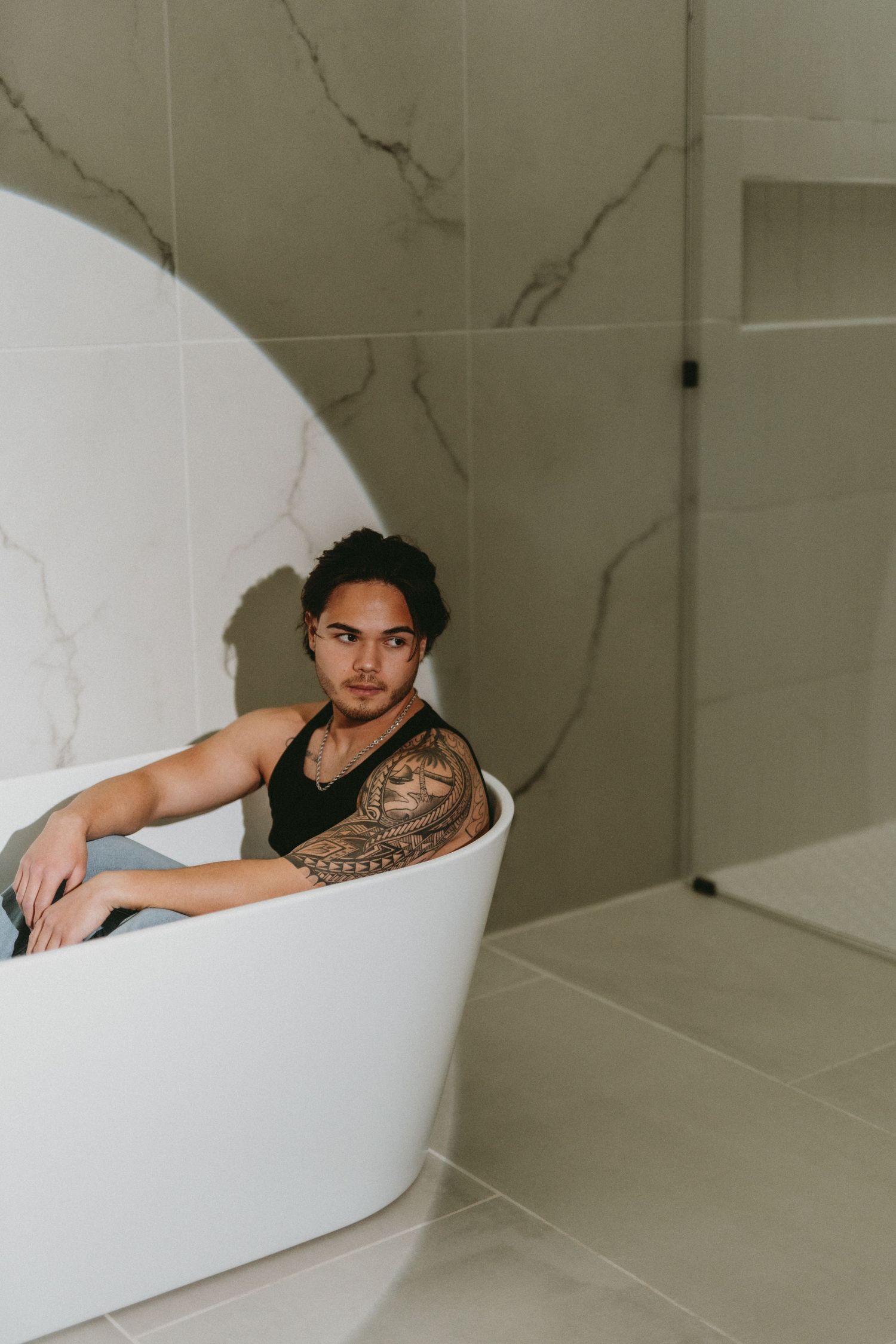 Tattooed man with Polynesian sleeve tattoo relaxing in modern freestanding bathtub in luxury marble bathroom.