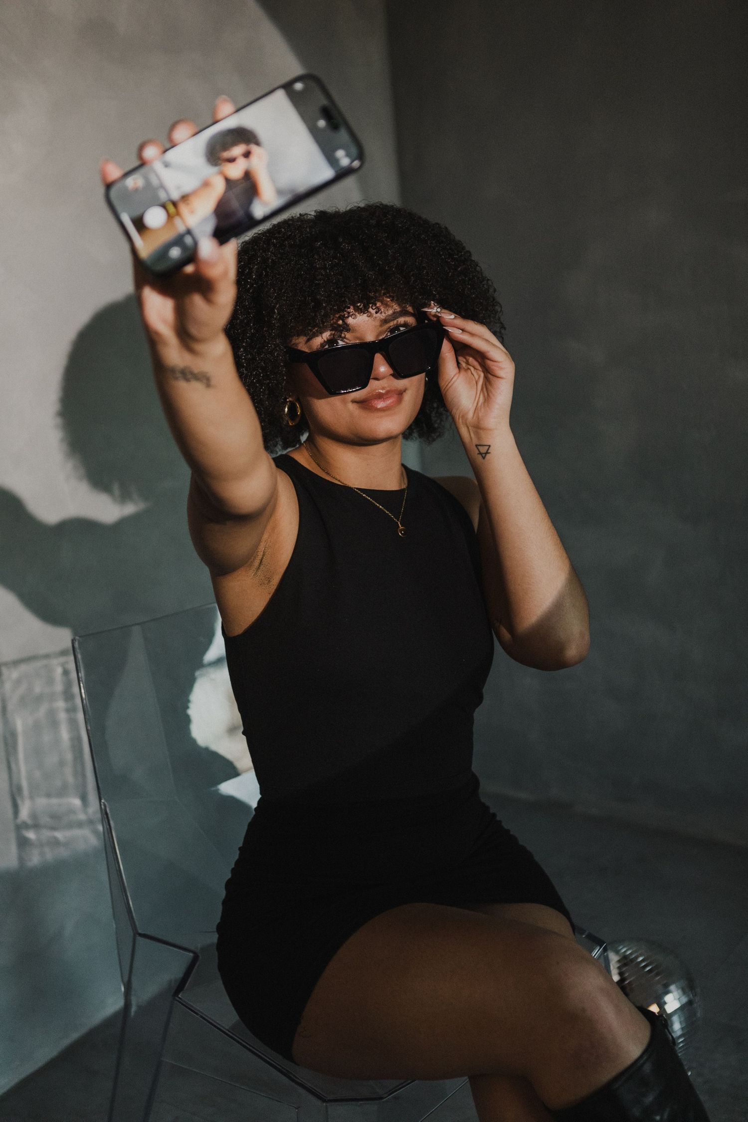 Stylish woman with curly hair in black dress takes selfie wearing sunglasses in gray studio.