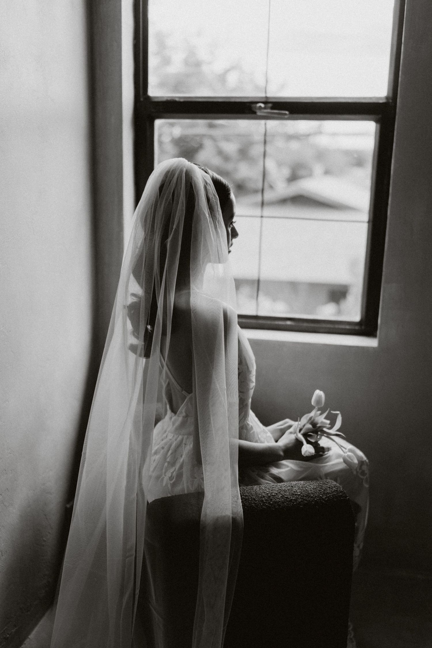 A black and white photo of a bride in a veil sitting by a window holding a flower.