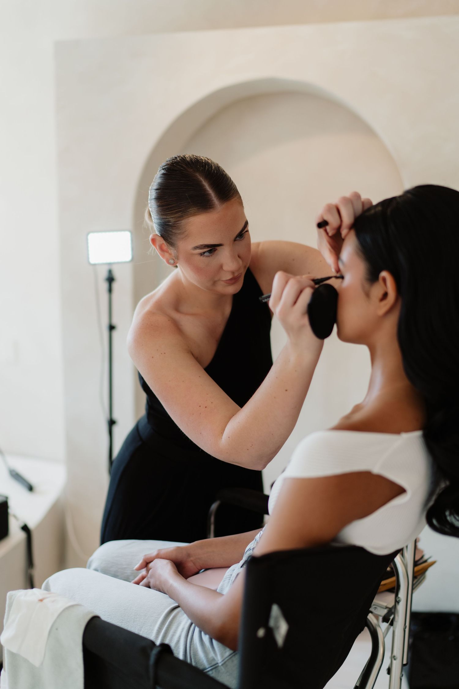 A makeup artist in black applies eye makeup to a bride in white during bridal preparation in a bright studio.
