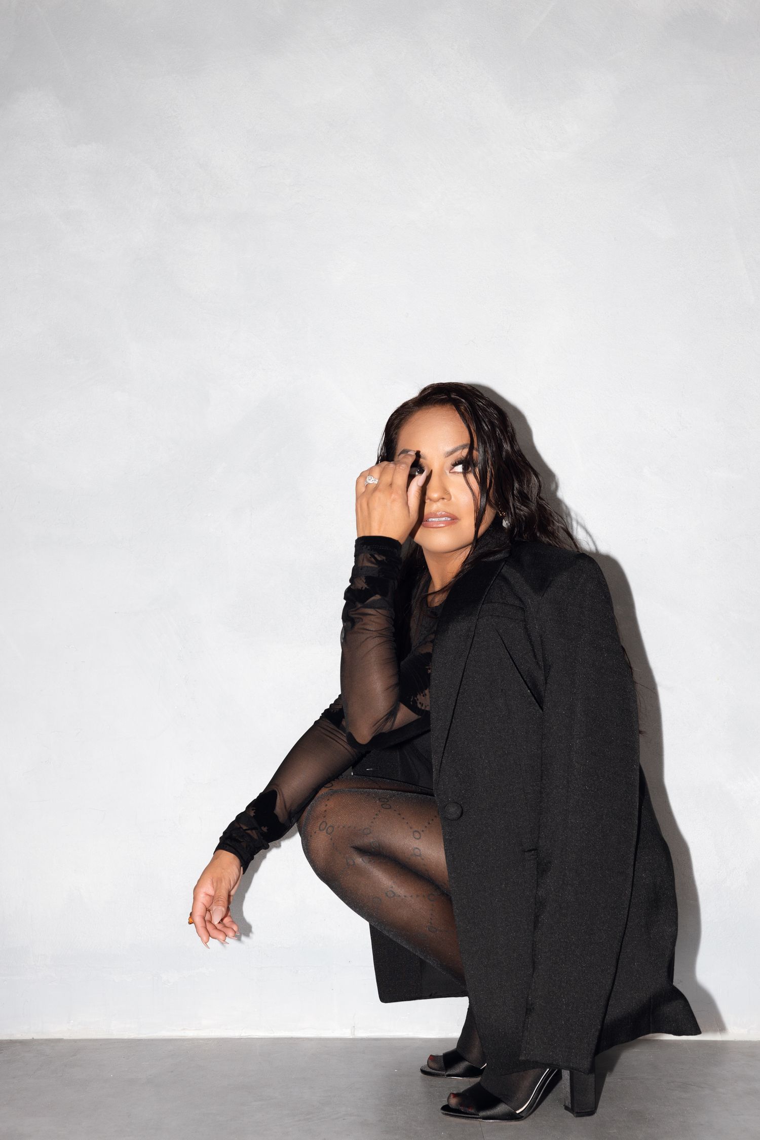 A woman in a black coat and sheer tights crouches against a white wall in a fashion pose.