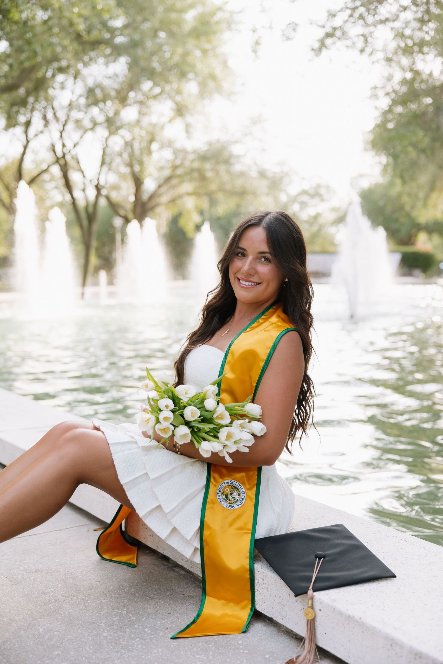 A University of South Florida grad sits by the fountain with lillies in her arms, by Tampa grad photographer Michaela Gom Photography.