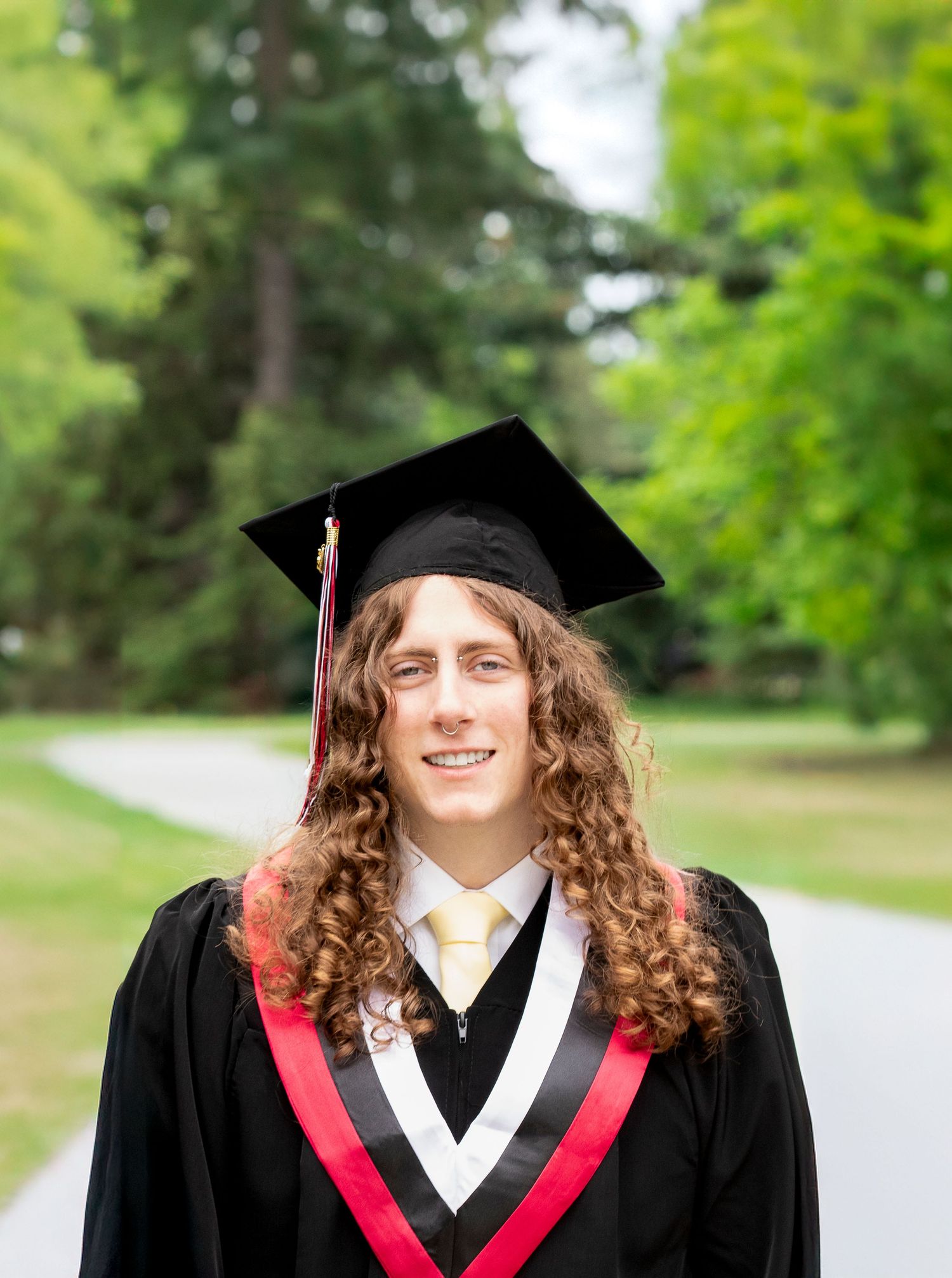 Graduate in traditional black cap and gown with red trim standing on outdoor path.