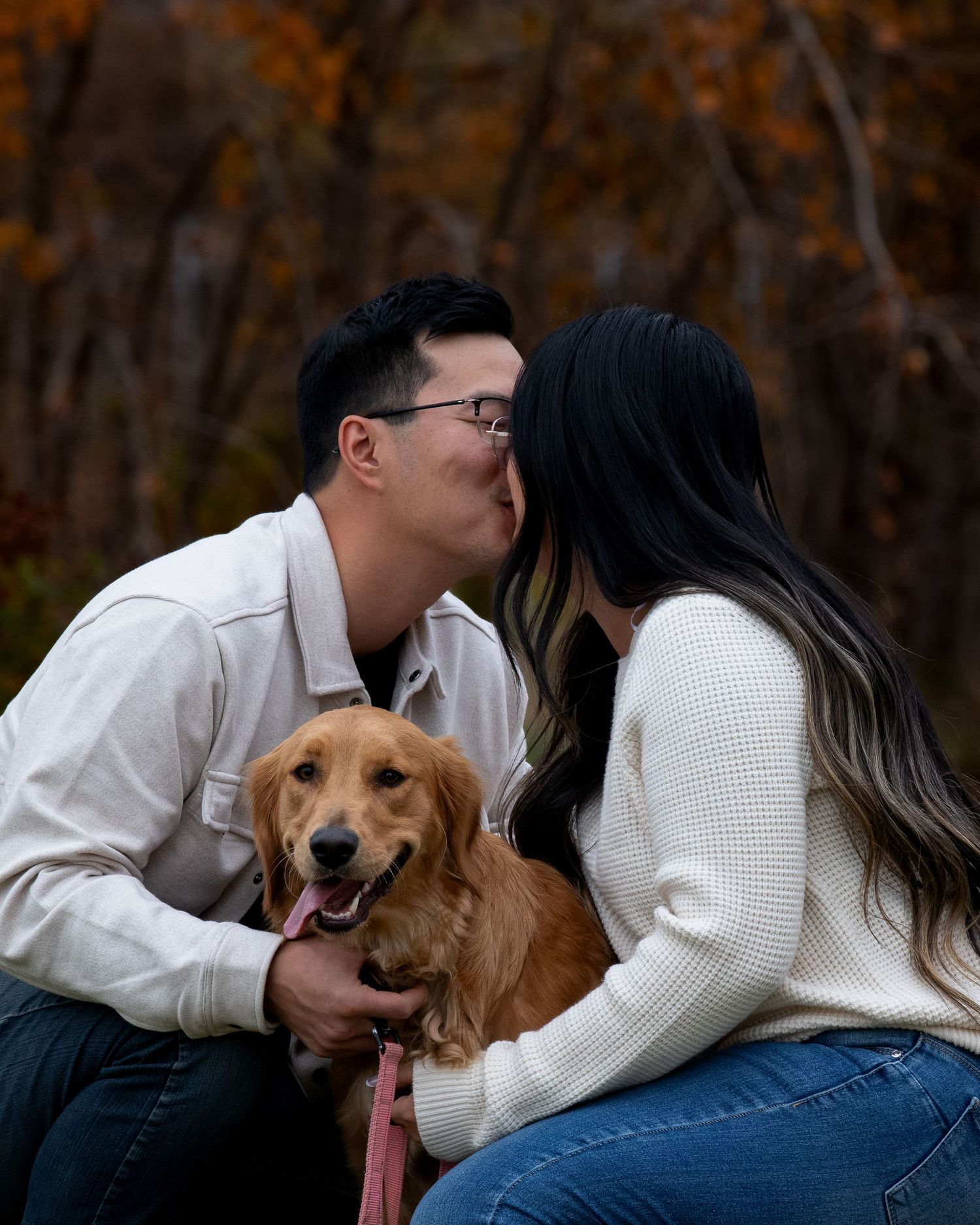 A happy golden retriever sitting between two people in a white sweater and jeans during autumn.