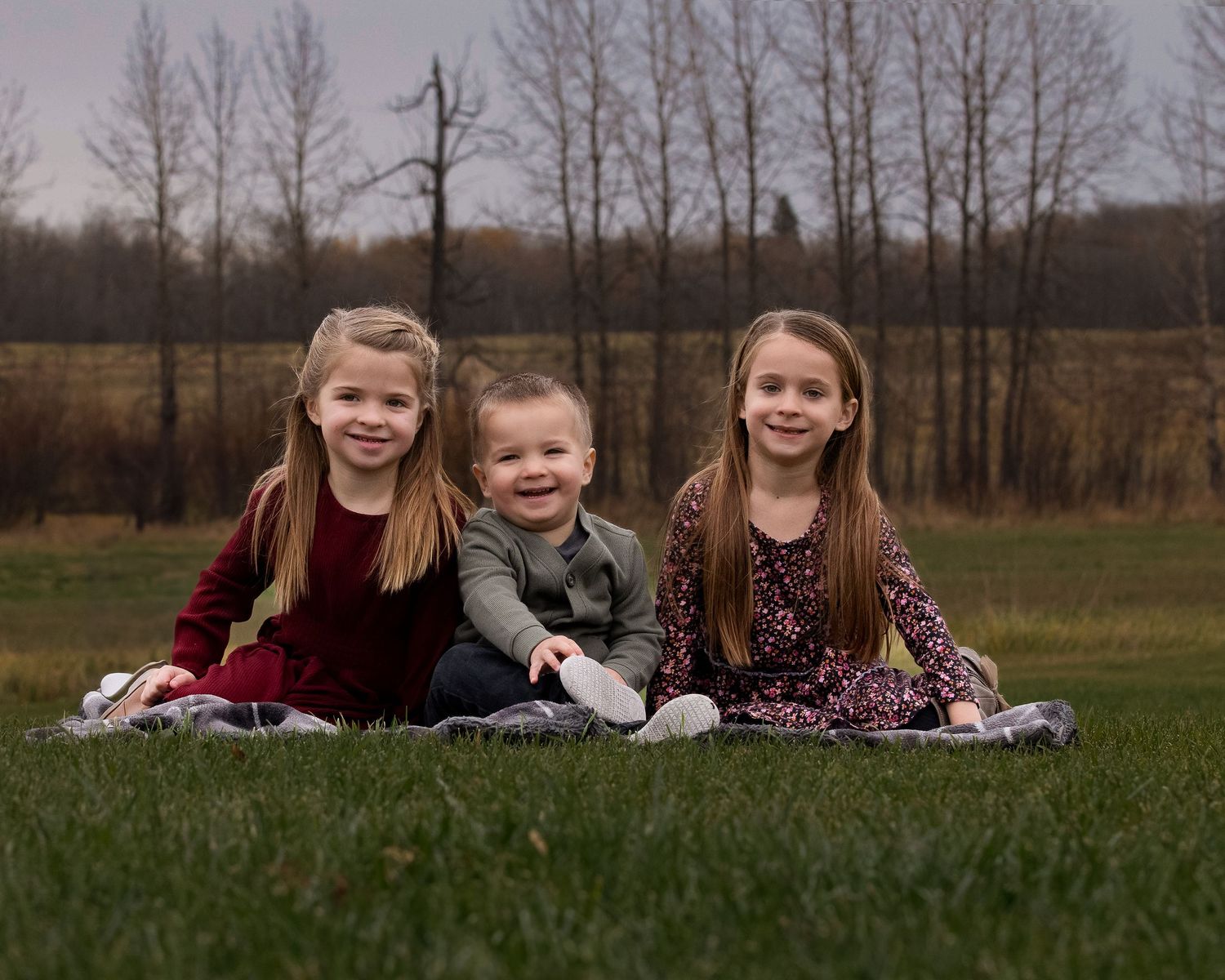 Three young children sit together on a blanket in a grassy field with bare trees in the background.